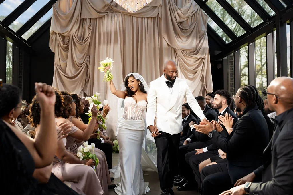 A bride and groom walking down the aisle after their wedding ceremony, surrounded by cheering guests in a bright, decorated venue with large windows.