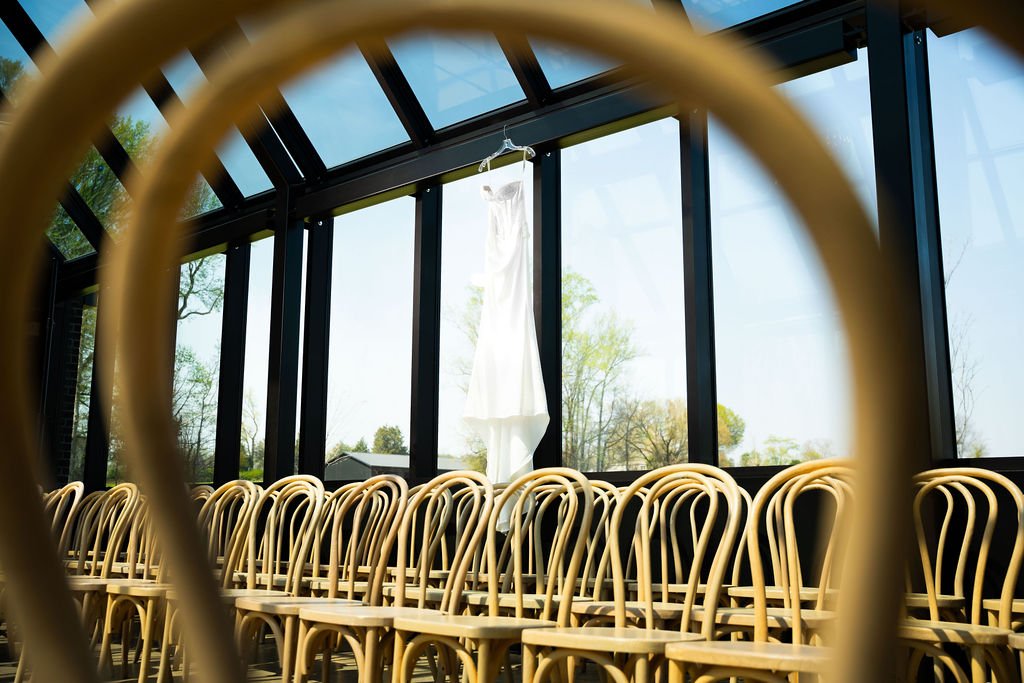 Wedding dress hanging in front of a large glass window with rows of chairs inside a sunlit conservatory or greenhouse setting.
