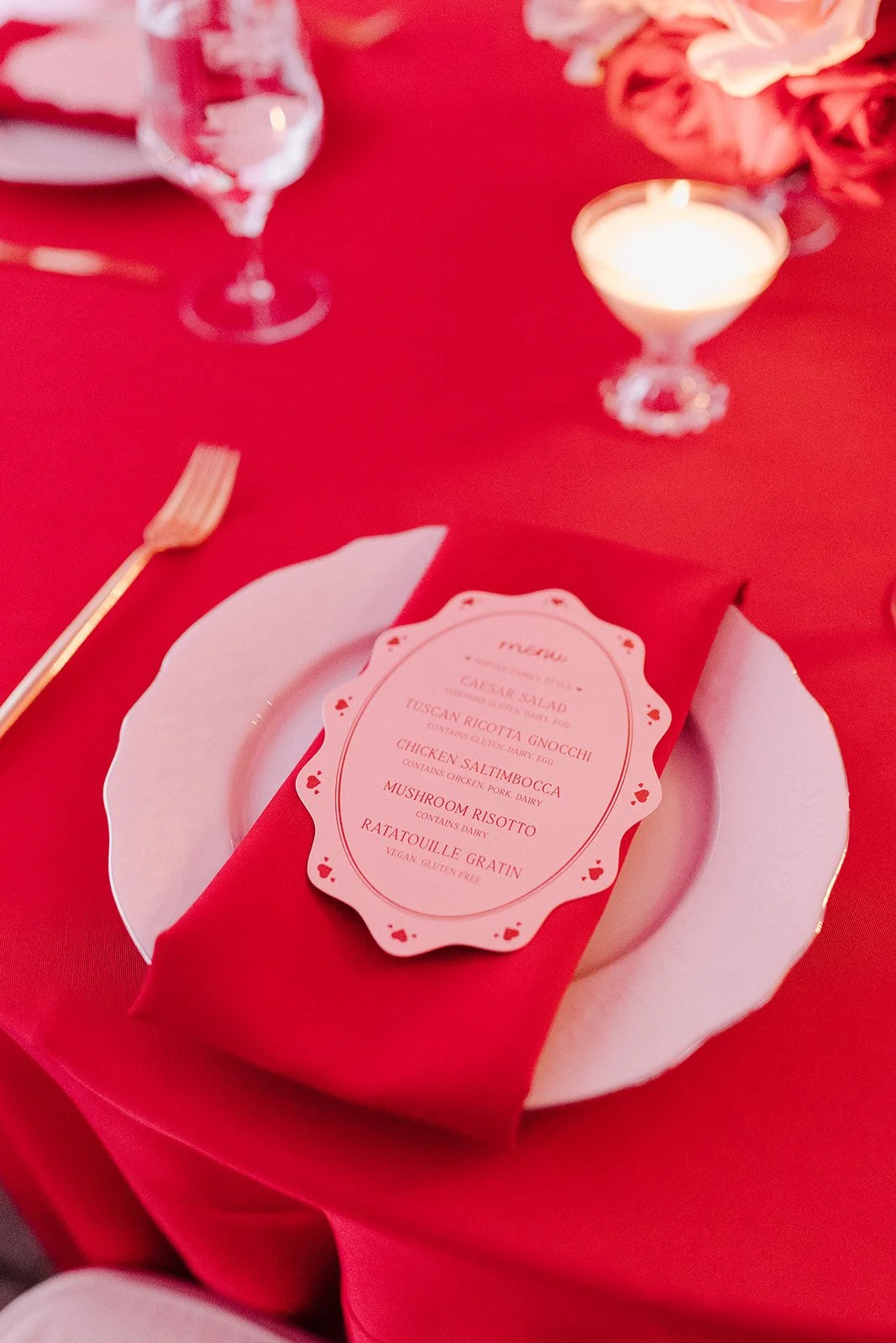 Table setting with a place card on a red napkin, a white plate, gold fork, glass of water, lit candle, and pink flowers in the background.