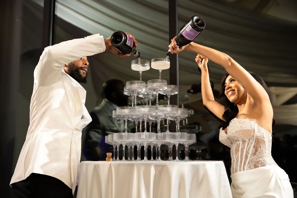A couple in wedding attire pours champagne into glasses on a champagne tower during a celebration.