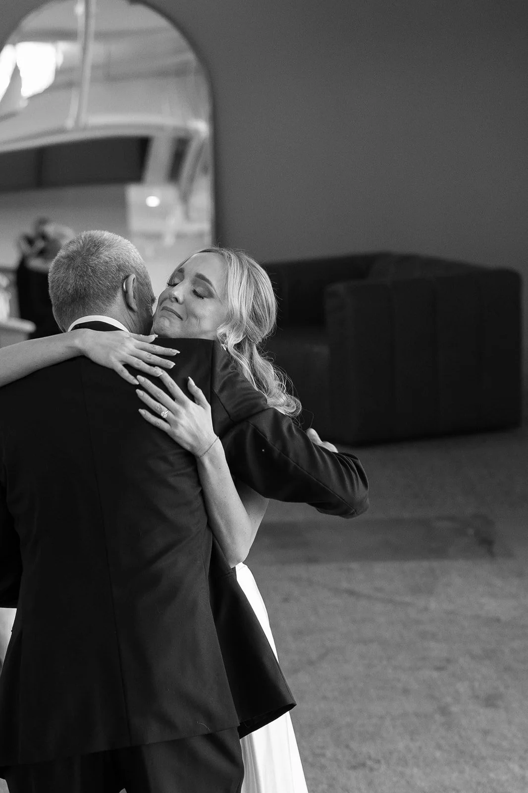 A woman with wavy blonde hair in a white dress embraces a man in a dark tuxedo, sharing a tender moment in a dance or hug.