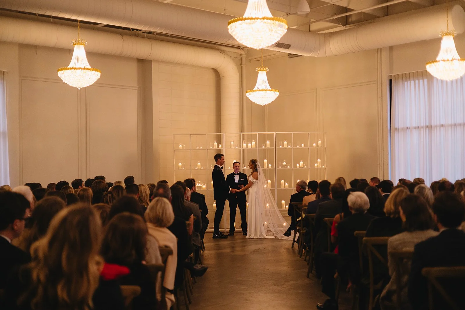 A wedding ceremony taking place indoors with the bride and groom exchanging vows in front of a seated audience. The background features several lit candles on a shelf, creating a warm and intimate atmosphere. The room is decorated with large chandeli