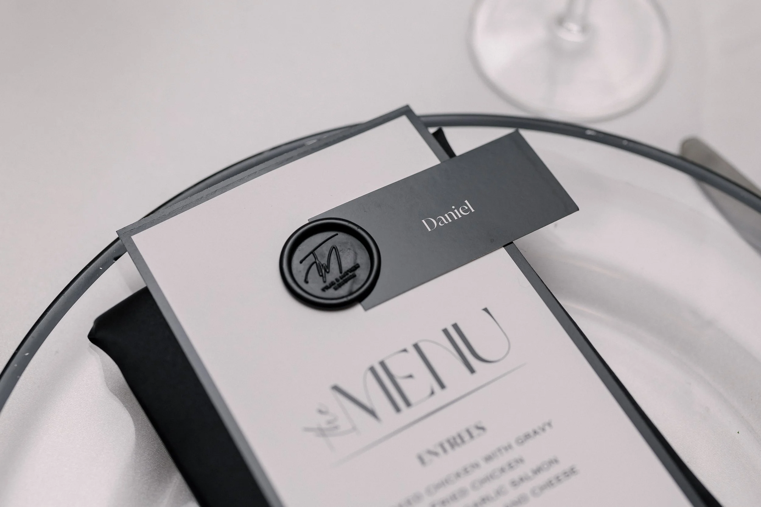 A close-up view of a menu with a black circular seal that has a stylized 'TD' logo and the name 'Daniel' in a gray rectangular card. A wine glass is visible in the top right corner.