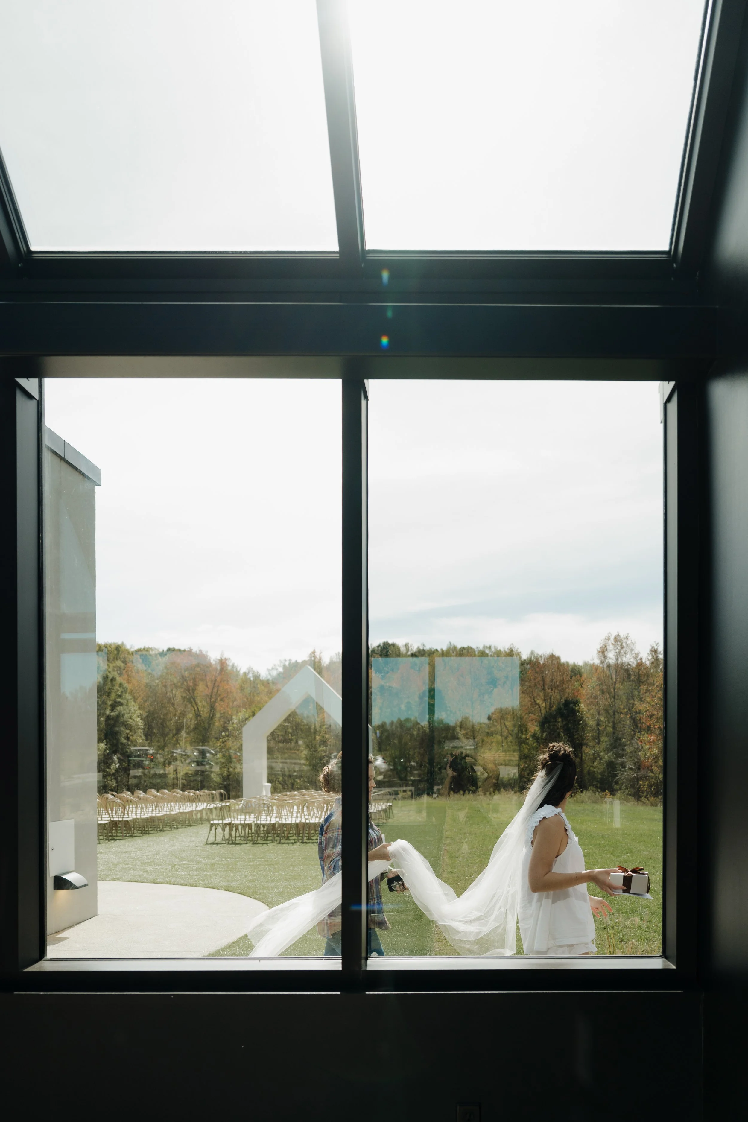 View through glass door showing two women, one in all white with a long dress, possibly a bride, and another in casual clothing with a plaid shirt, outside in a grassy area with chairs arranged in rows, and a backdrop of trees and overcast sky.