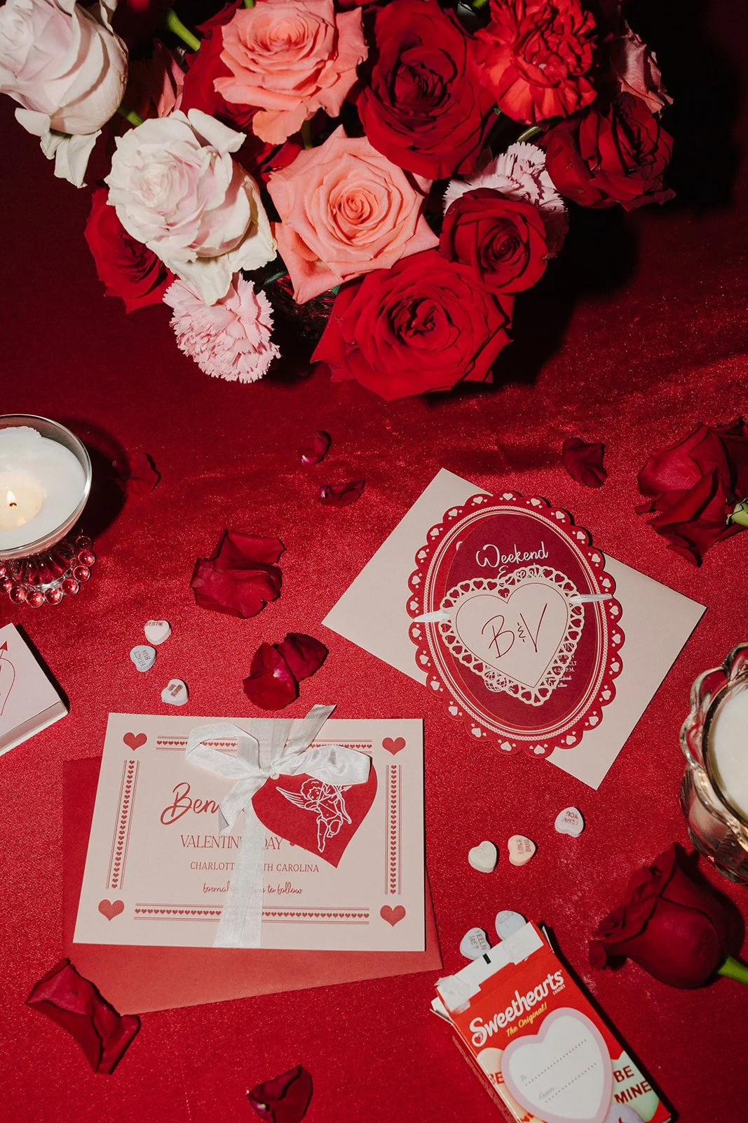 A Valentine's Day table setup with pink and red roses, scattered rose petals, a candle, heart-shaped confetti, a Valentine's Day card with a cupid illustration and text, and a box of Heart-shaped Sweethearts candies.