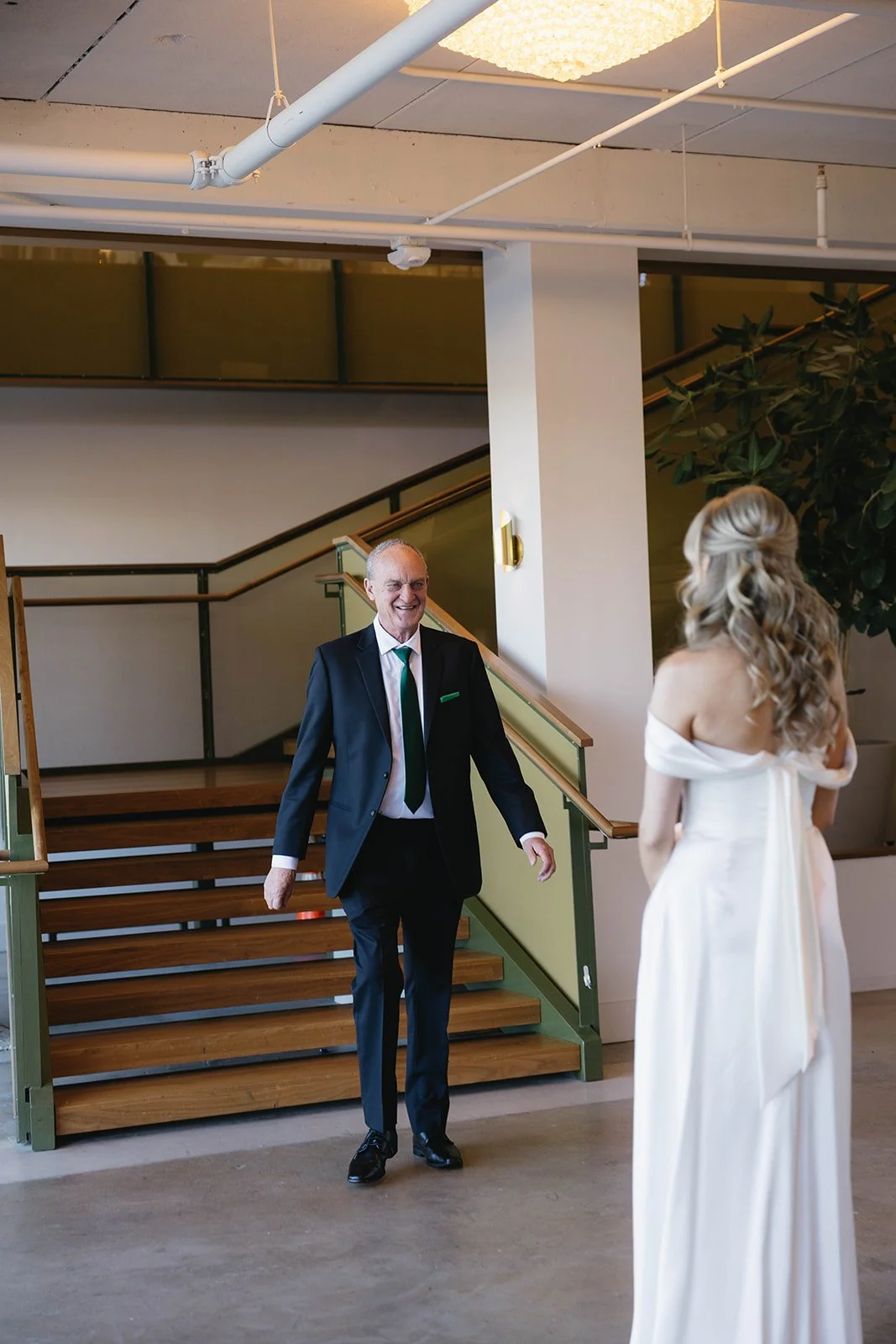 An older man in a dark suit smiling and approaching a woman in a white off-shoulder dress inside a building with wooden stairs and large green plant in the background.