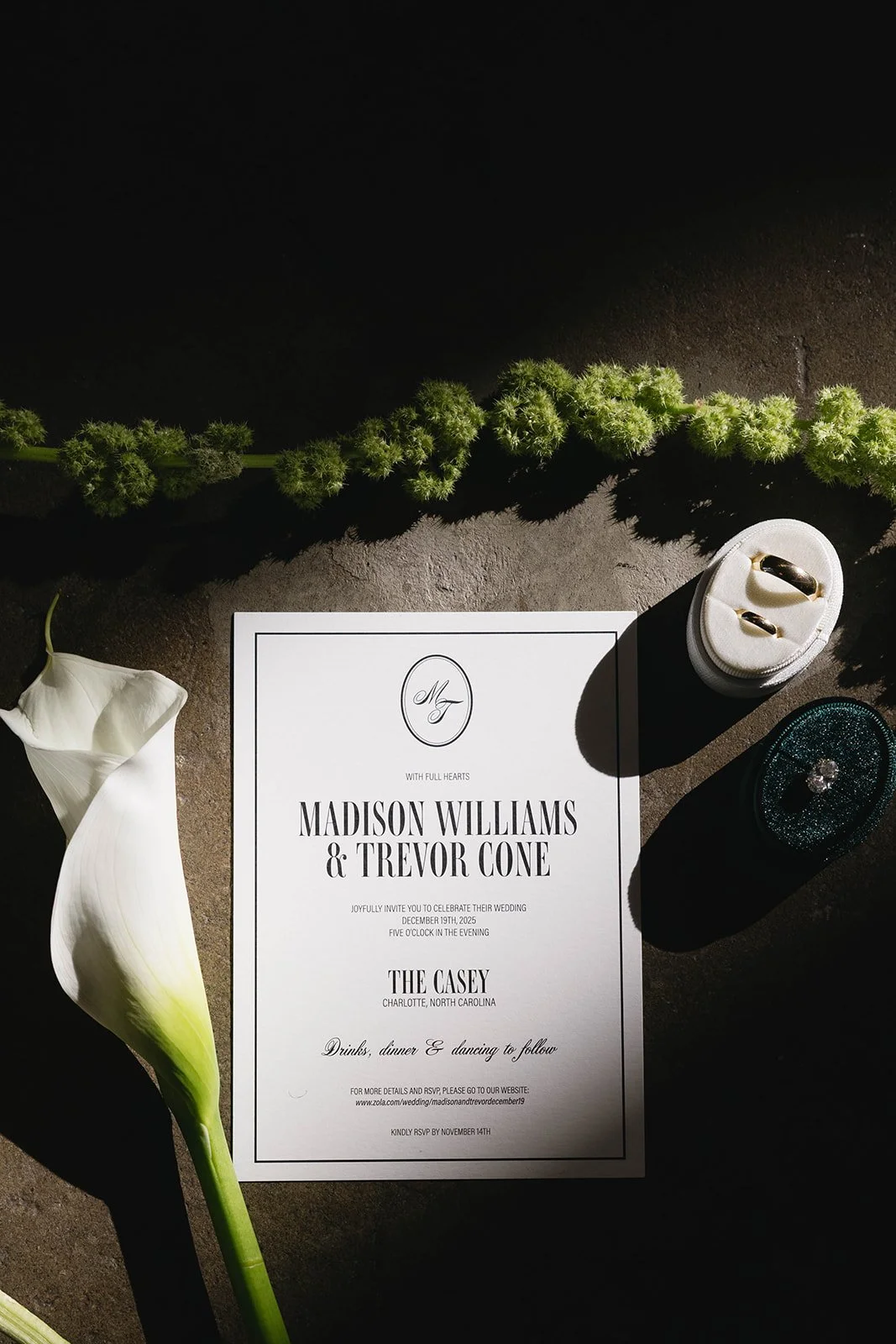 Wedding invitation with greenery, a calla lily flower, and jewelry on a dark surface.