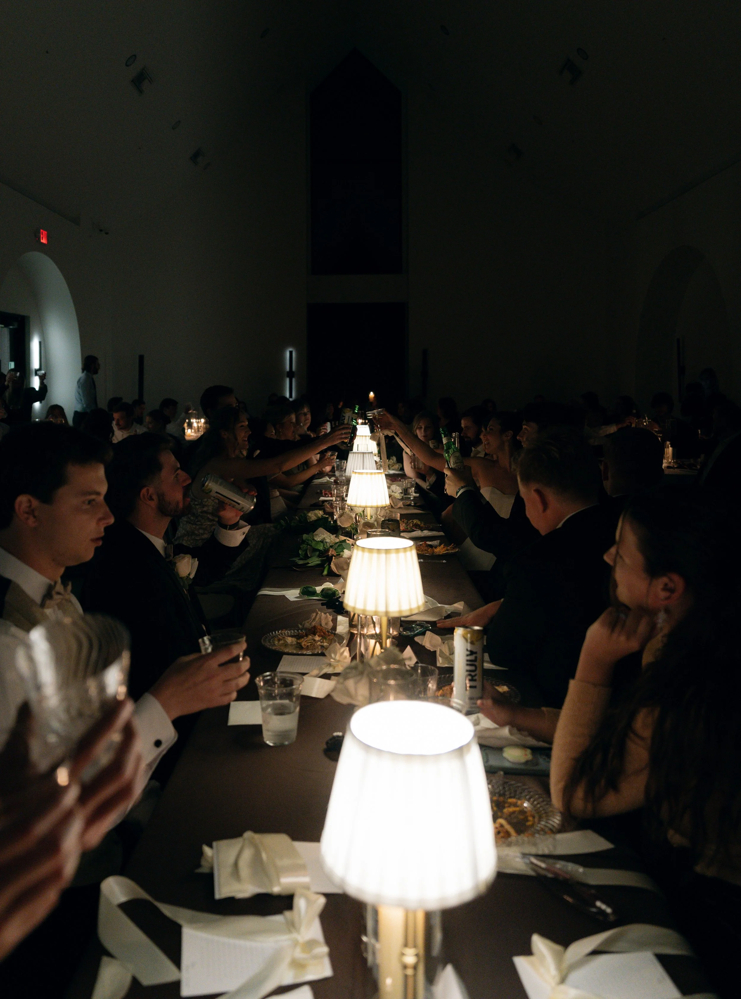 People sitting at a long dinner table in a dimly lit room, raising glasses in a toast, with table lamps casting light on their faces, and plates of food and napkins scattered across the table.