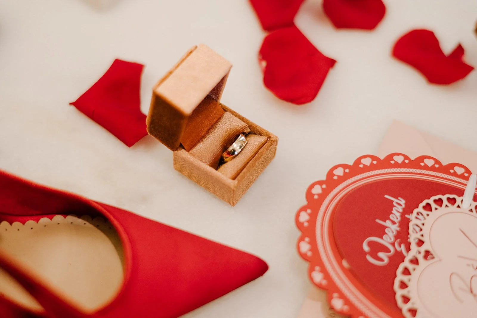 A small ring box with a silver ring inside, surrounded by red rose petals, a heart-shaped box, and a Valentine's Day card.