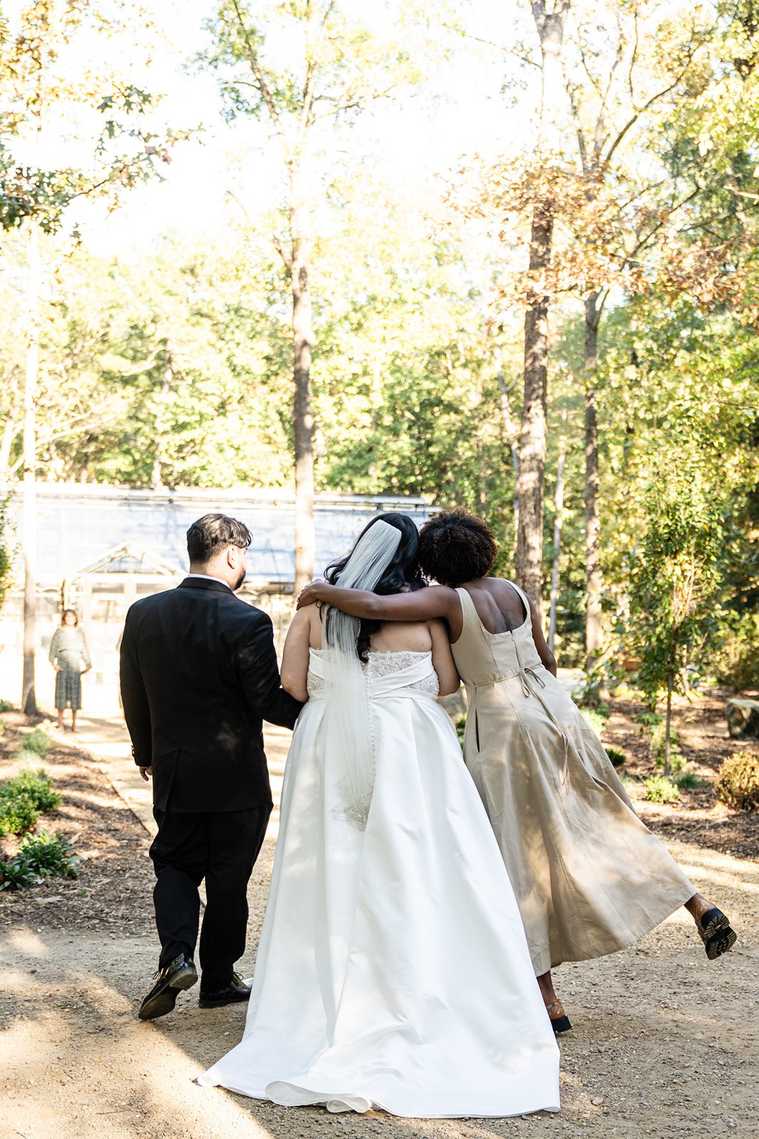 A wedding scene with a bride, groom, and another woman embracing outdoors on a sunny day, with trees and a woman in the background.