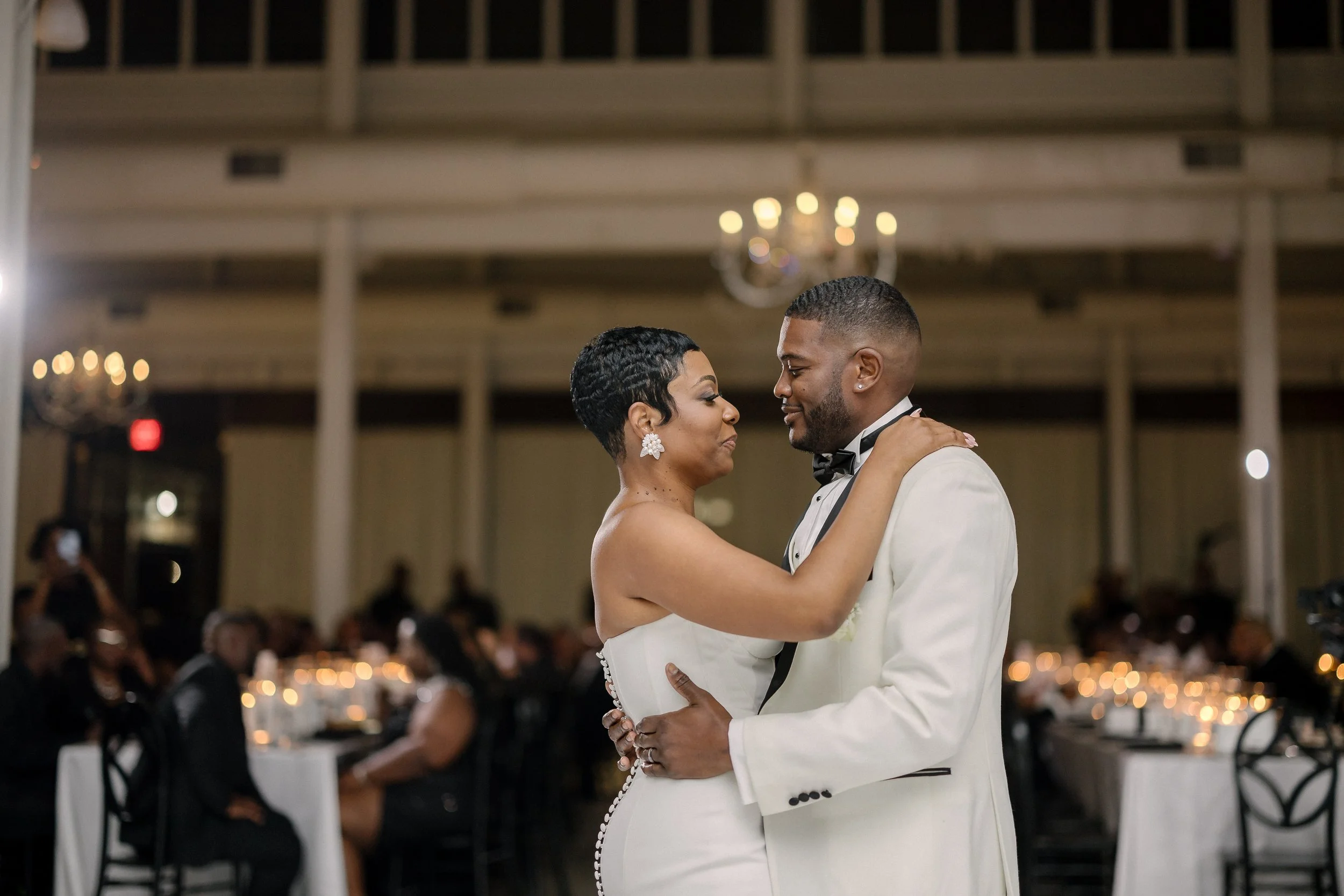 A couple dancing closely at a formal event with many seated guests in the background, elegant lighting, and chandeliers in a banquet hall.