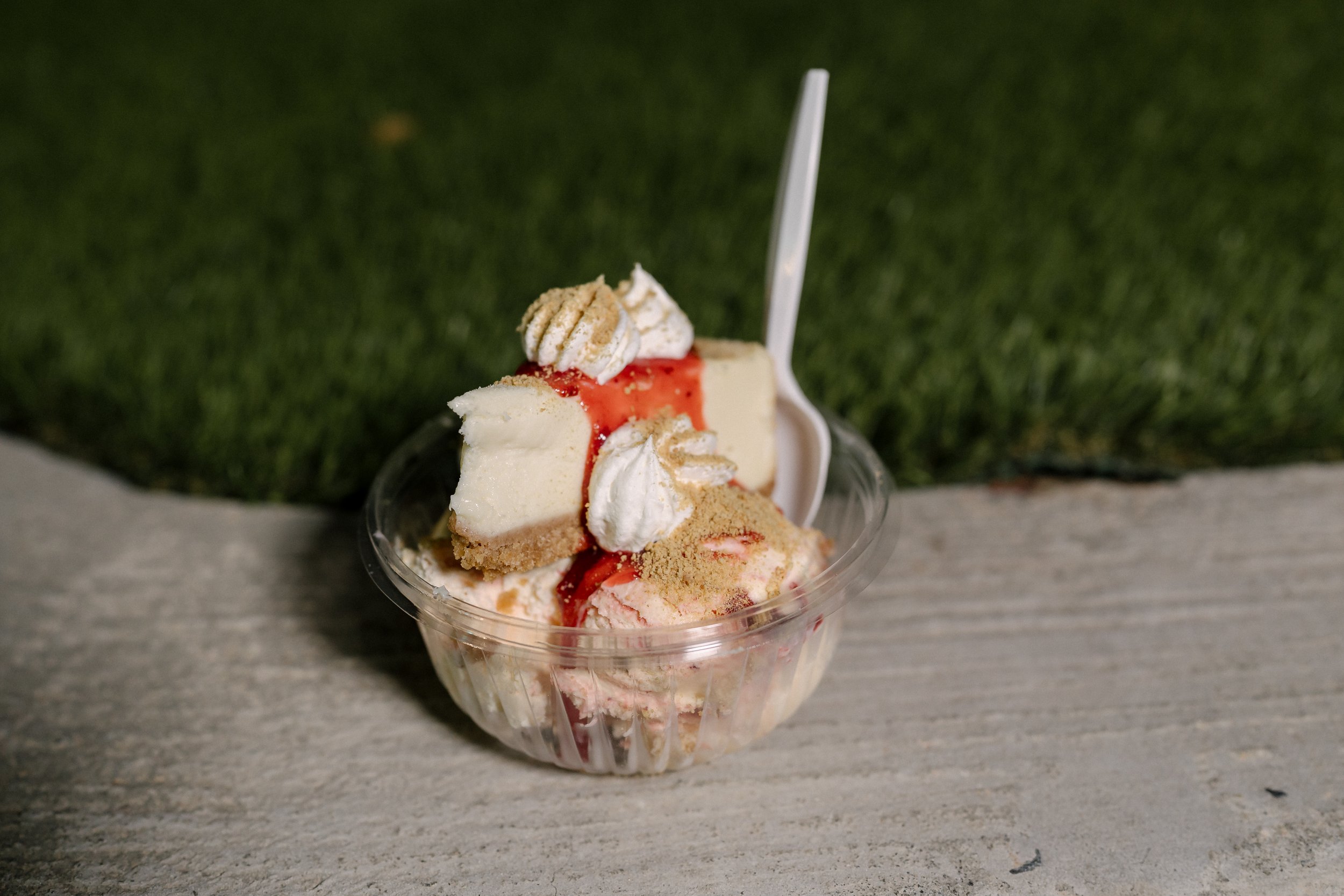 A small glass bowl of vanilla ice cream topped with red syrup, dollops of whipped cream, and crumbled cookie pieces, with a plastic spoon inserted.