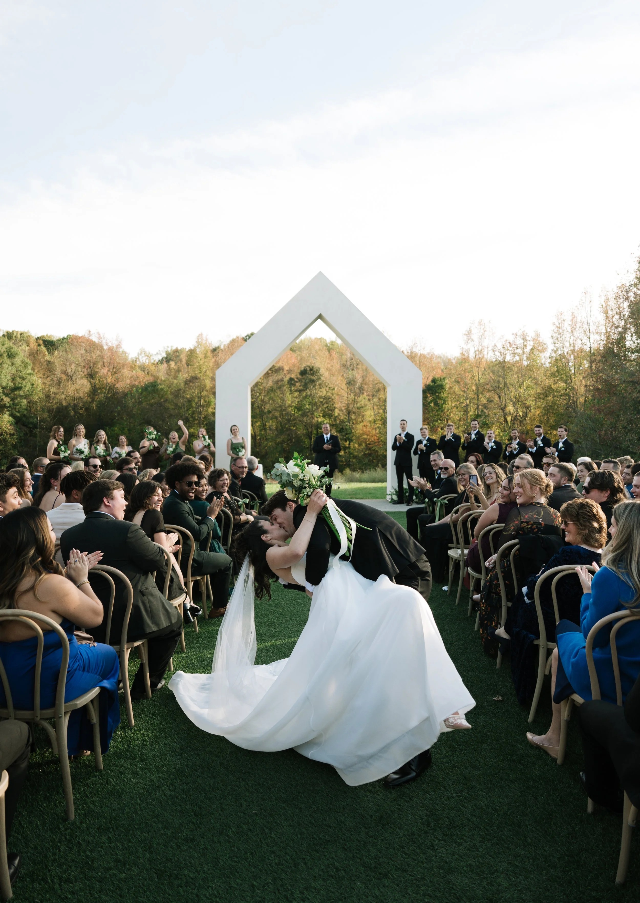 A wedding ceremony outdoors with the bride and groom performing a kiss, surrounded by guests seated on chairs on green grass. There is a white arch and a forest background.