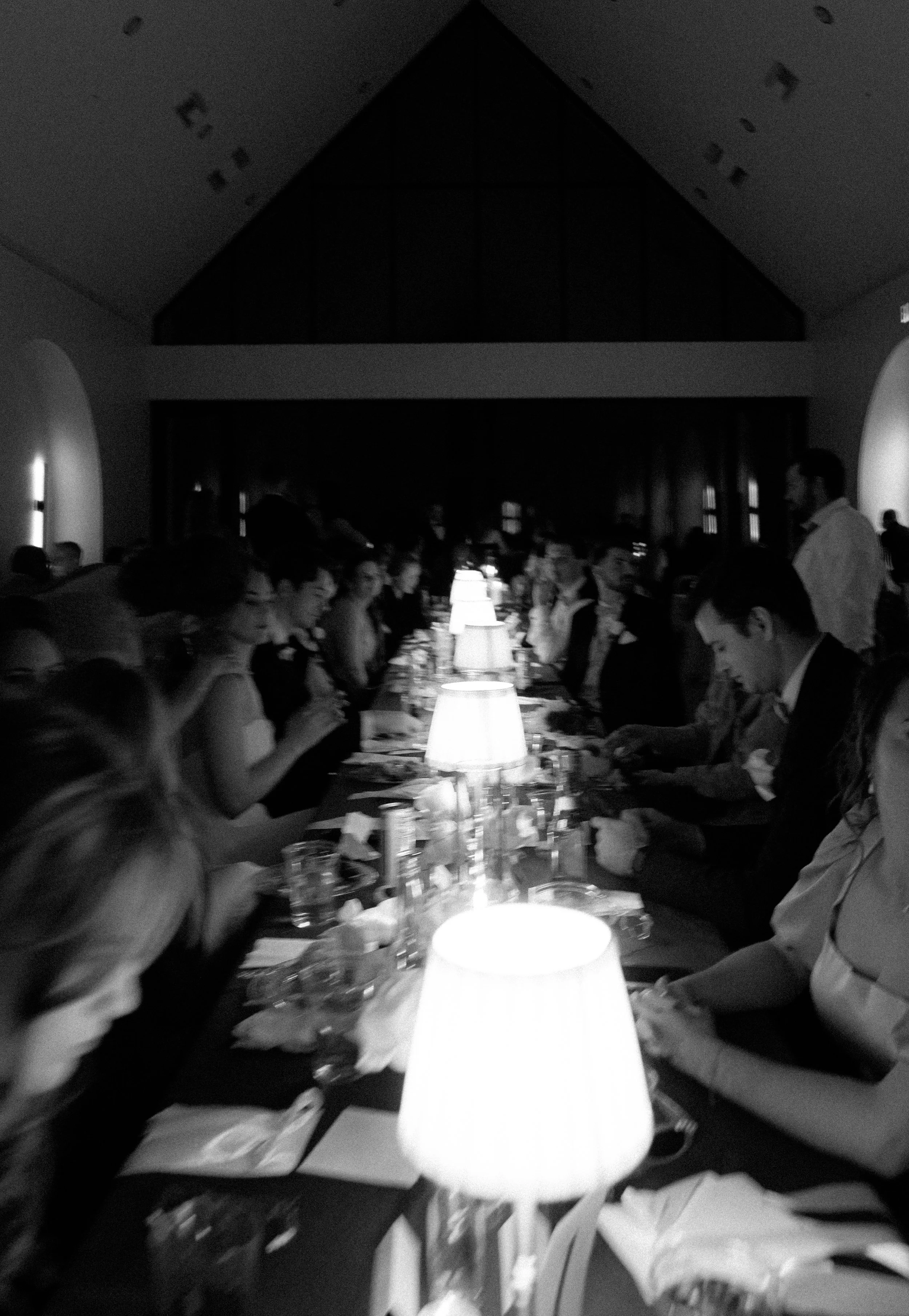 People sitting at a long dining table in a dimly lit room with lamps, engaged in conversation and dining.