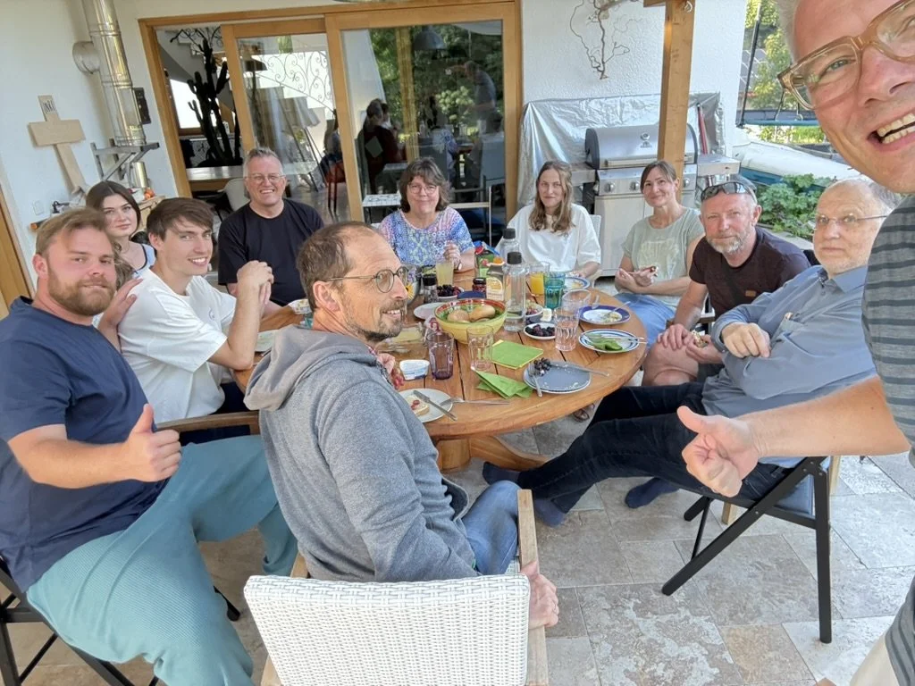 Gruppe von elf Menschen sitzen und sitzen um einen runden Holztisch beim Essen im Freien auf einer Terrasse, mit Lebensmitteln, Tellern und Getränken auf dem Tisch.