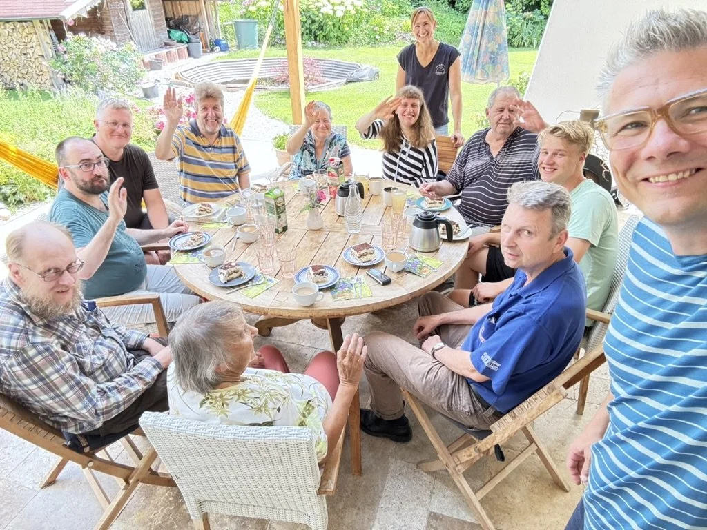 Gruppe von Menschen beim Frühstück im Garten, lachend und winkend, mit Tischen, Kaffee, Kuchen, und em Spielplatz im Hintergrund.