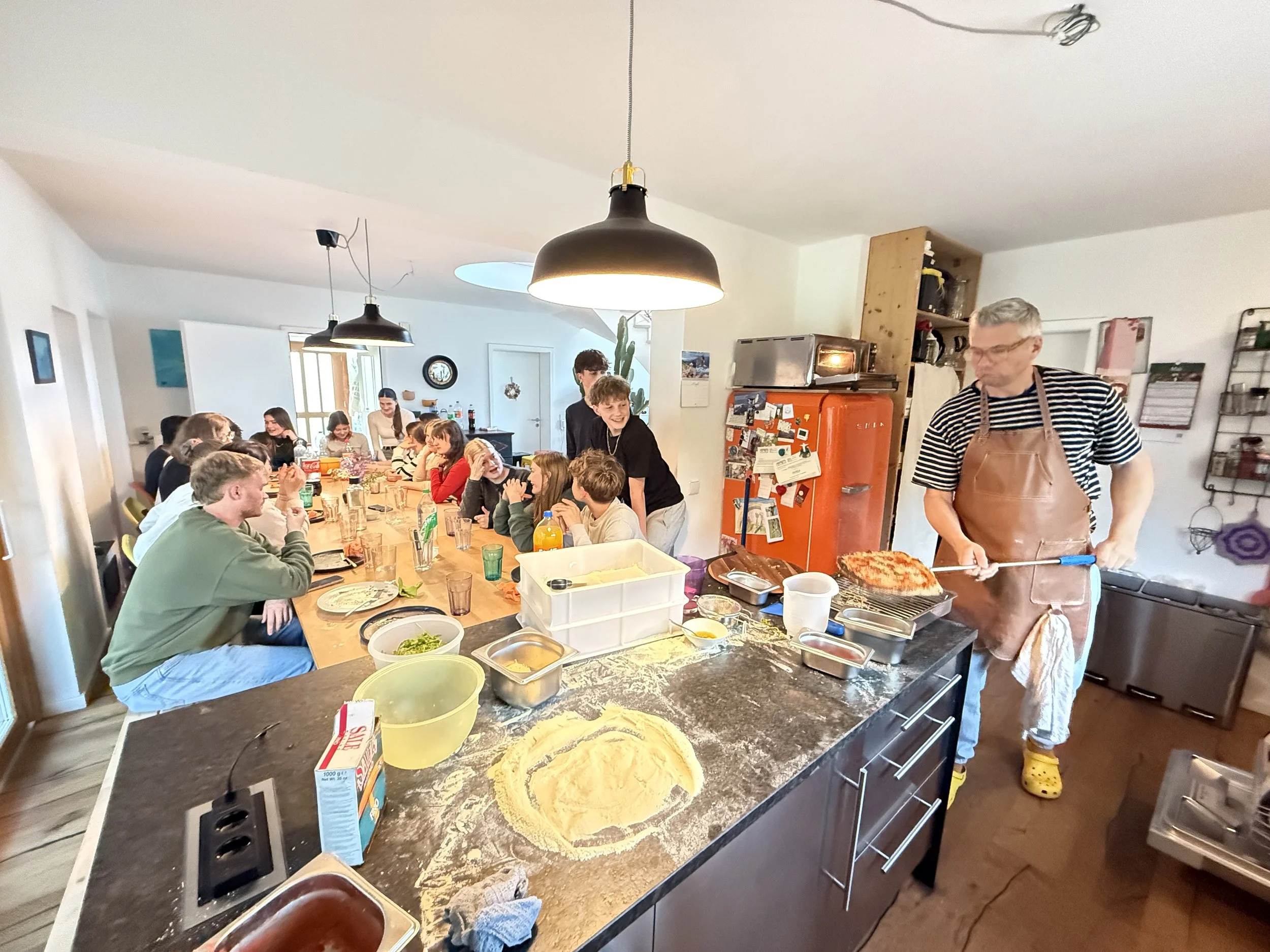 Eine Gruppe von Menschen sitzt an einem langen Esstisch in einer offenen Küche und unterhält sich, während ein Mann eine Pizza auf einem Küchenbrett schneidet. Im Vordergrund sind Mehl, Töpfe und Küchenutensilien auf einer Kücheninsel sichtbar.