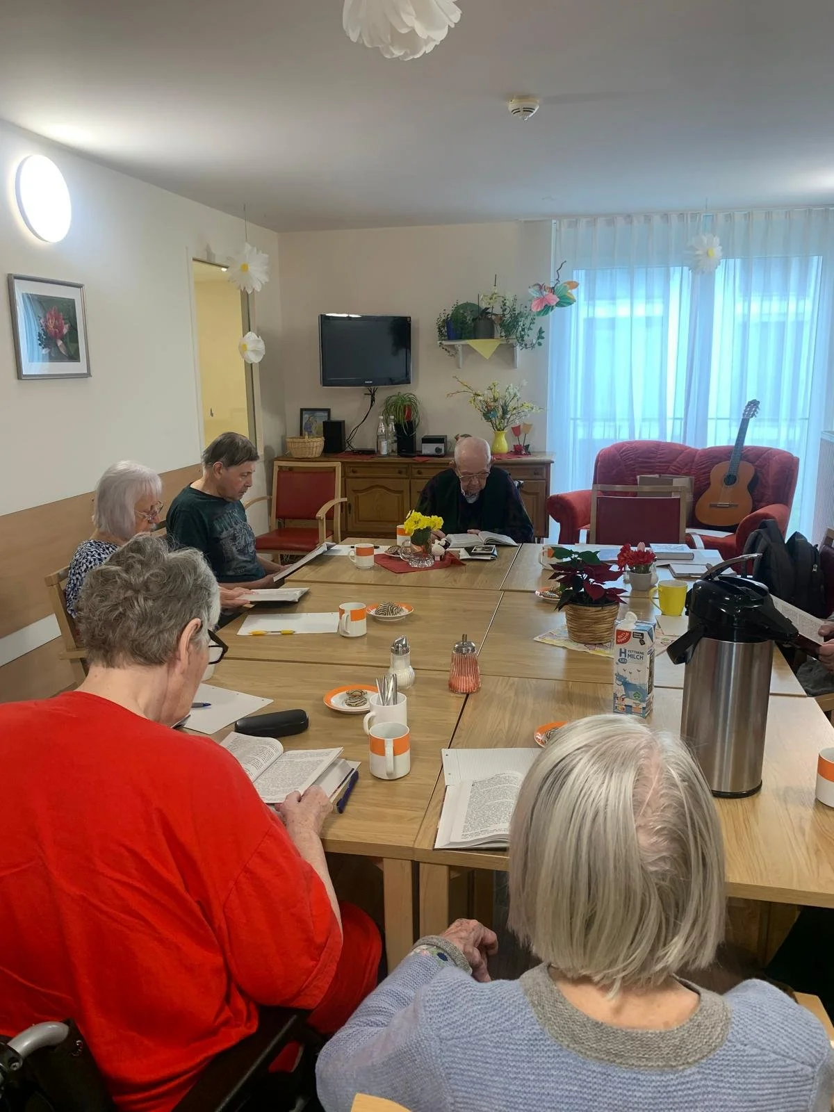 Mehrgenerationen-Treffen in einem gemütlichen Raum mit einem großen Holztablett, auf dem Kaffee, Kekse, Zeitschriften und Schreibutensilien liegen. Im Hintergrund sind Bücher, Pflanzen, eine Gitarre, ein Fernseher und ein Sofa mit Kissen zu sehen.