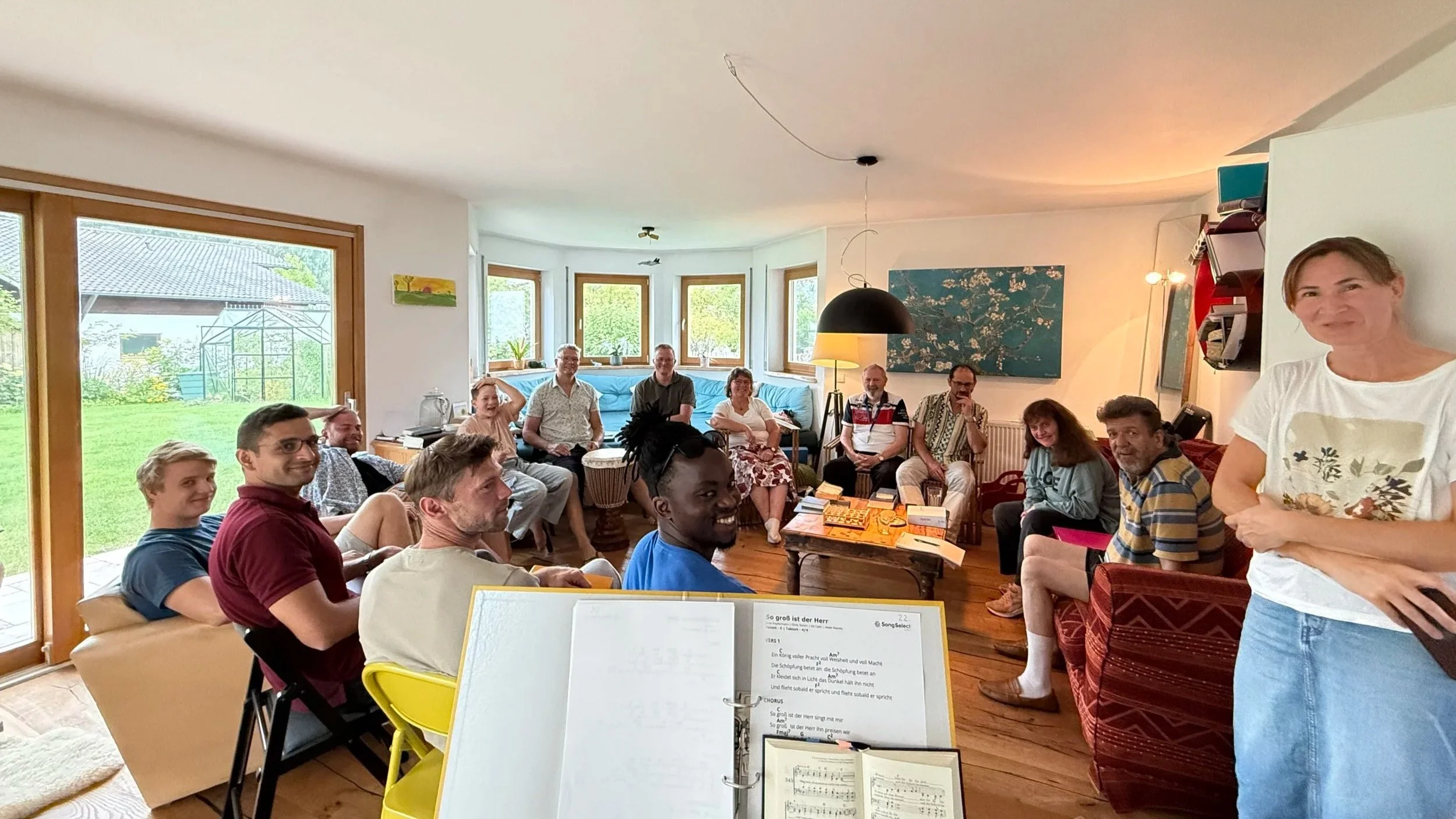 Menschen sitzen in einem Wohnzimmer bei einem Treffen. Einige sehen in die Kamera, die meisten schauen auf den Musikordner auf dem Tisch. Es steht ein Musiker vor der Gruppe, die auf einer roten Couch sitzt, die vor einer großen Fensterfront mit Blick auf einen Garten steht.