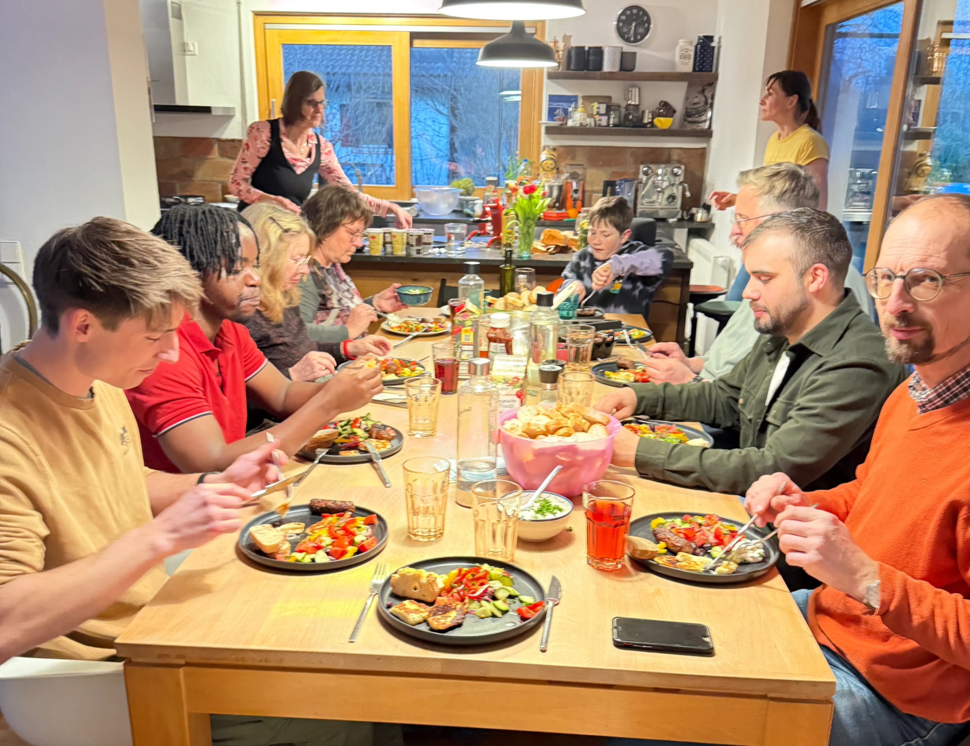 Eine Gruppe von Menschen sitzt an einem Esstisch und isst gemeinsam. Im Hintergrund sieht man eine Küche mit Fenstern und mehreren Personen, die sich dort beschäftigen.