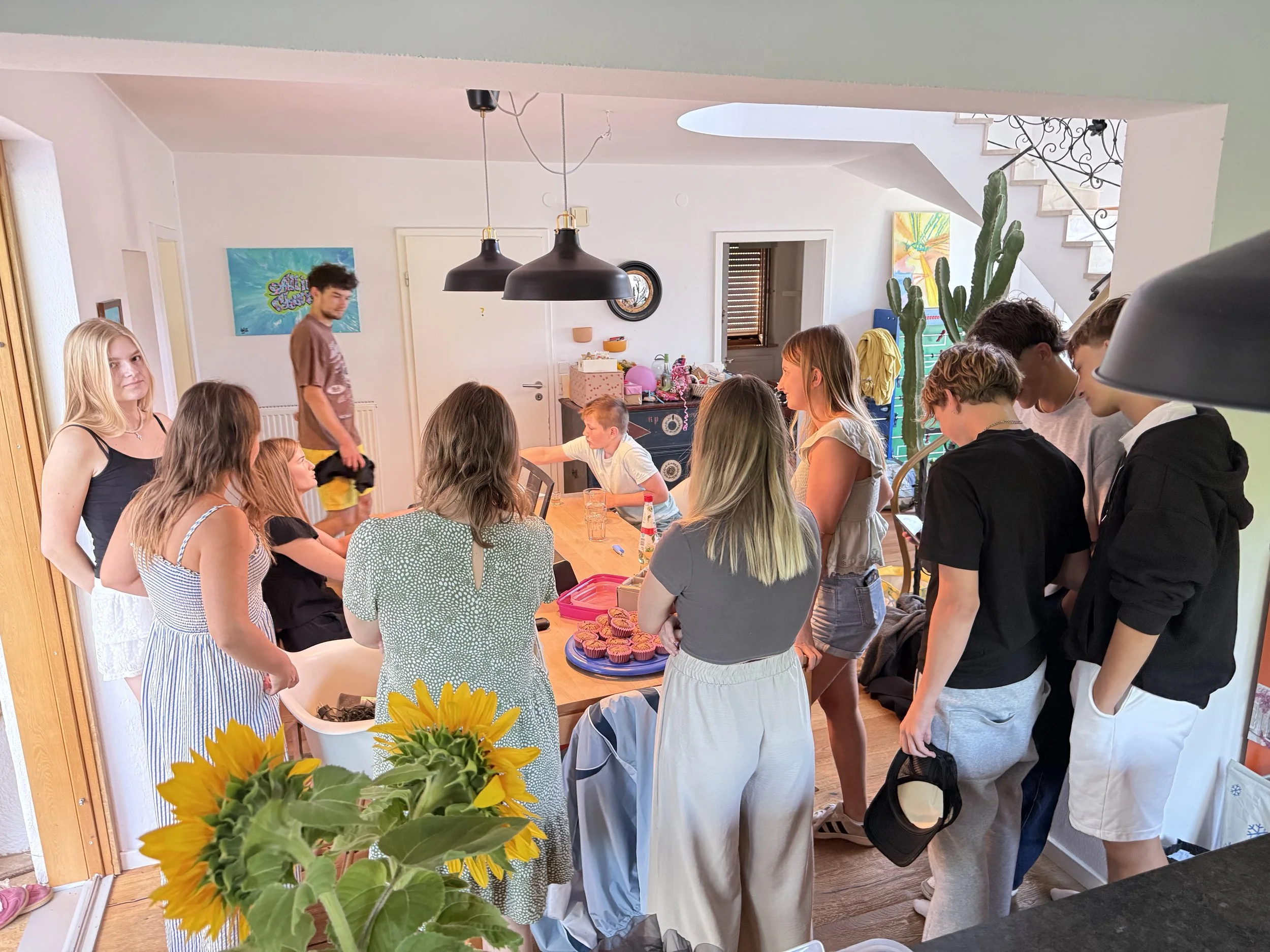 Mehrere Jugendliche versammeln sich in einer hellen Küche, einige sitzen um einen Tisch mit Kuchen und Snacks, andere stehen und sprechen, im Hintergrund eine Treppe und ein großer Kaktus.