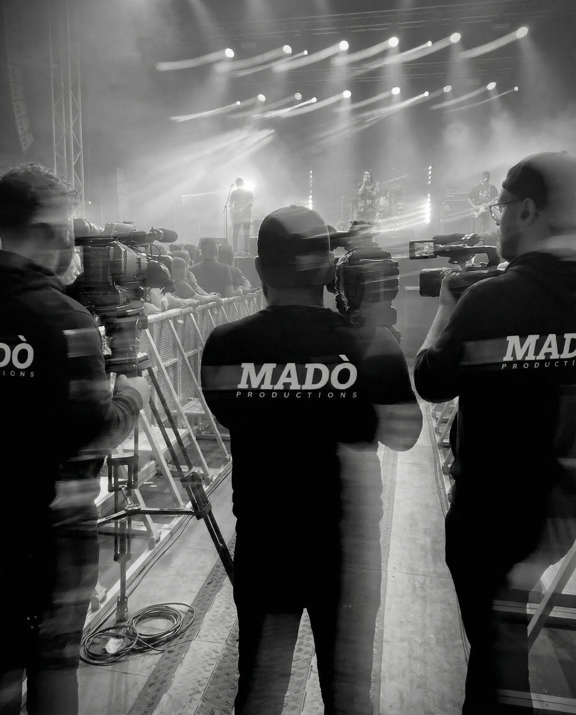 Tre cameramen con magliette con scritto 'MADÒ PRODUCTIONS' stanno filmando un concerto dal backstage, con pubblico e band sul palco illuminato da luci lampeggianti.