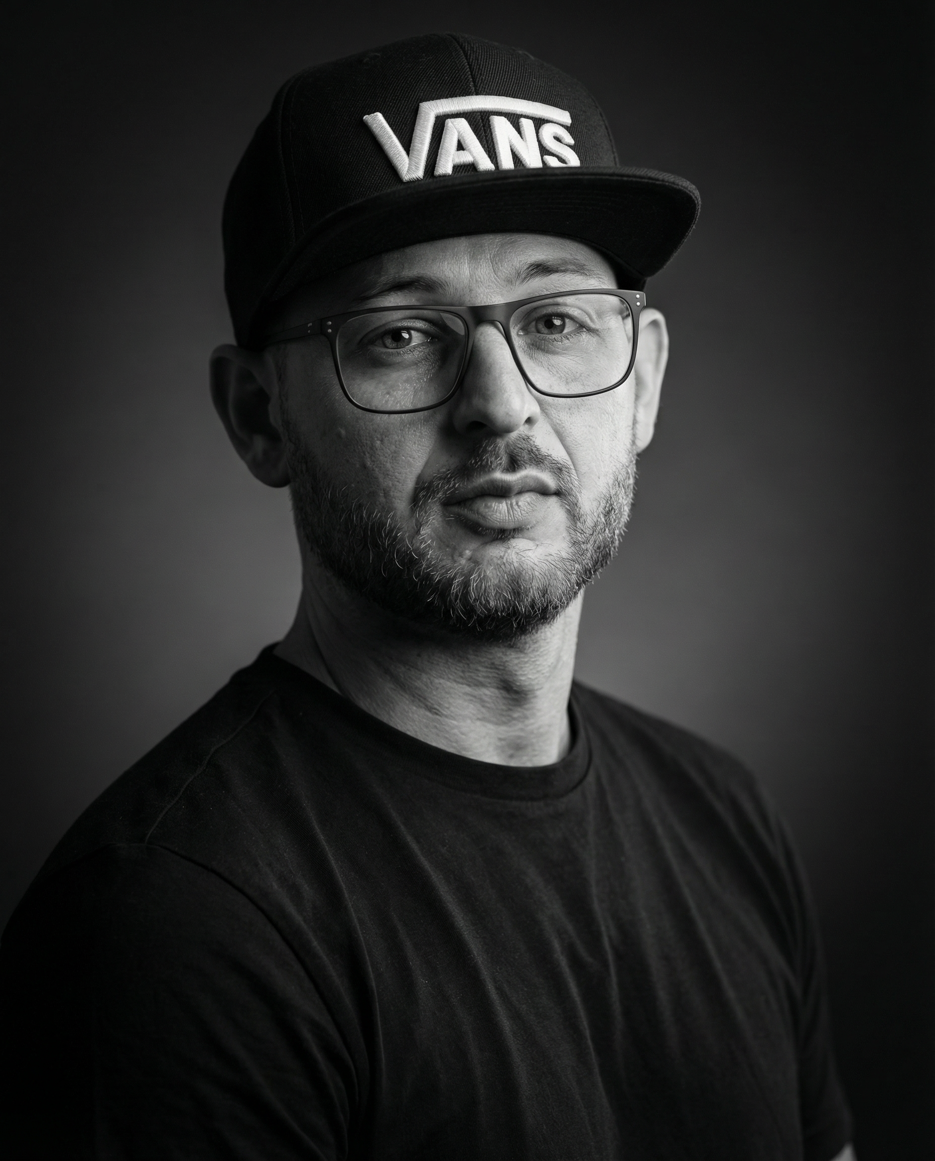 Ritratto in bianco e nero di un uomo con cappello e occhiali, con barba e sorriso leggero, sfondo scuro.