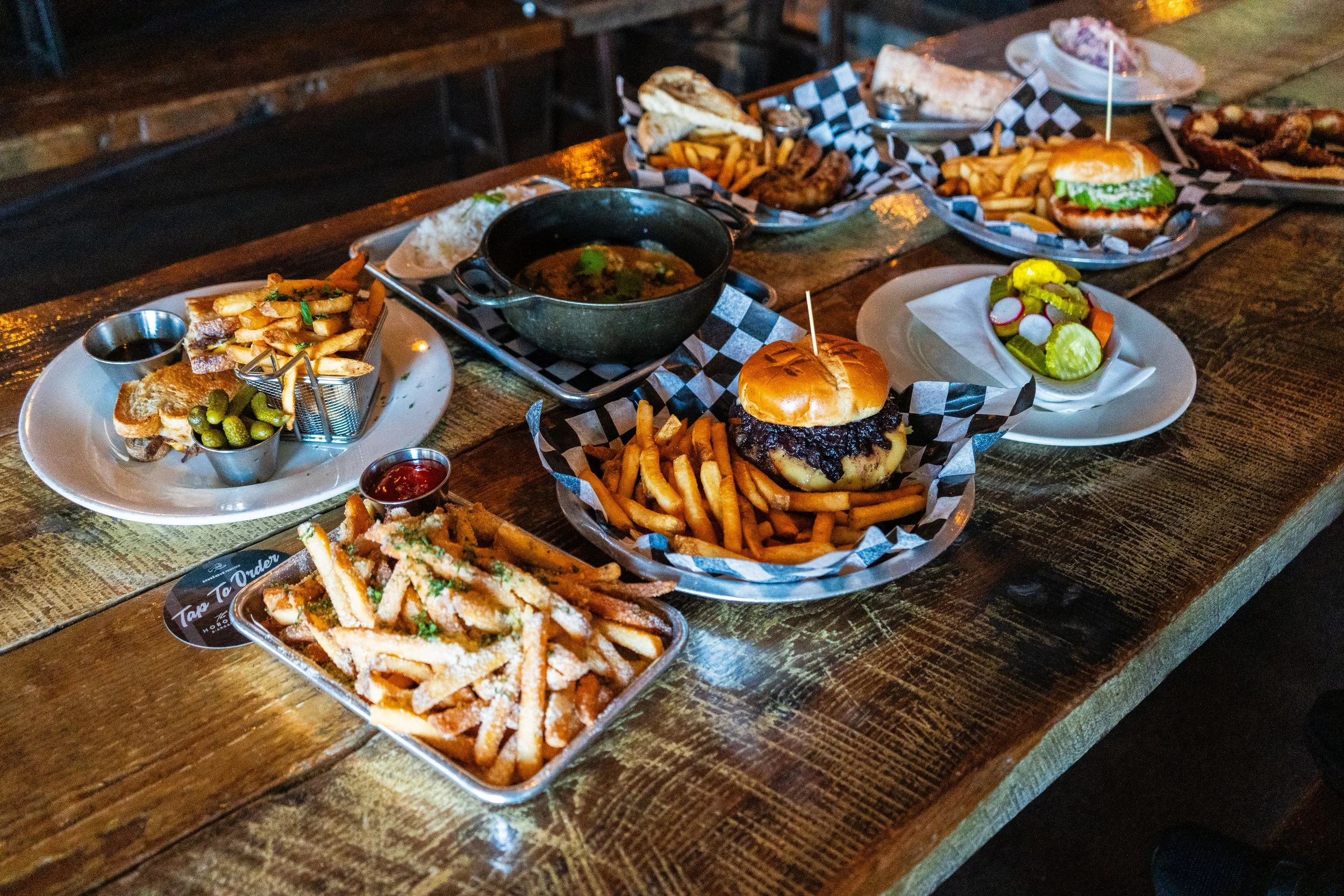 Variety of grilled sandwiches, burgers, and fries on a rustic wooden table. Dishes include pickles, a bowl of soup, and dipping sauces.