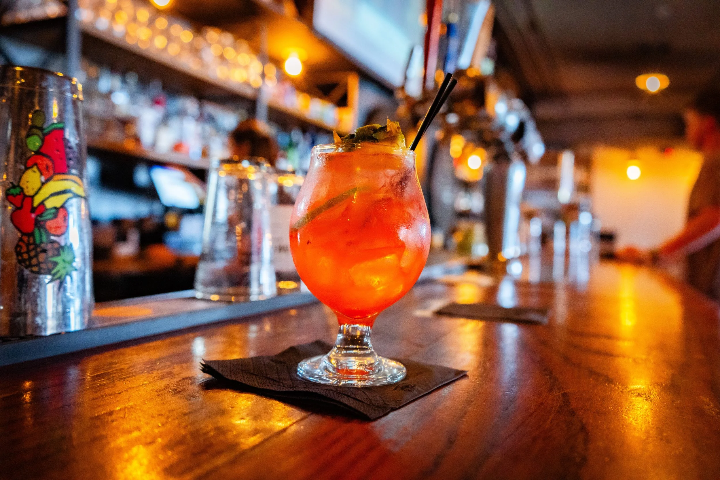 Colorful cocktail in a glass on a bar counter with blurred background of bottles and glasses.