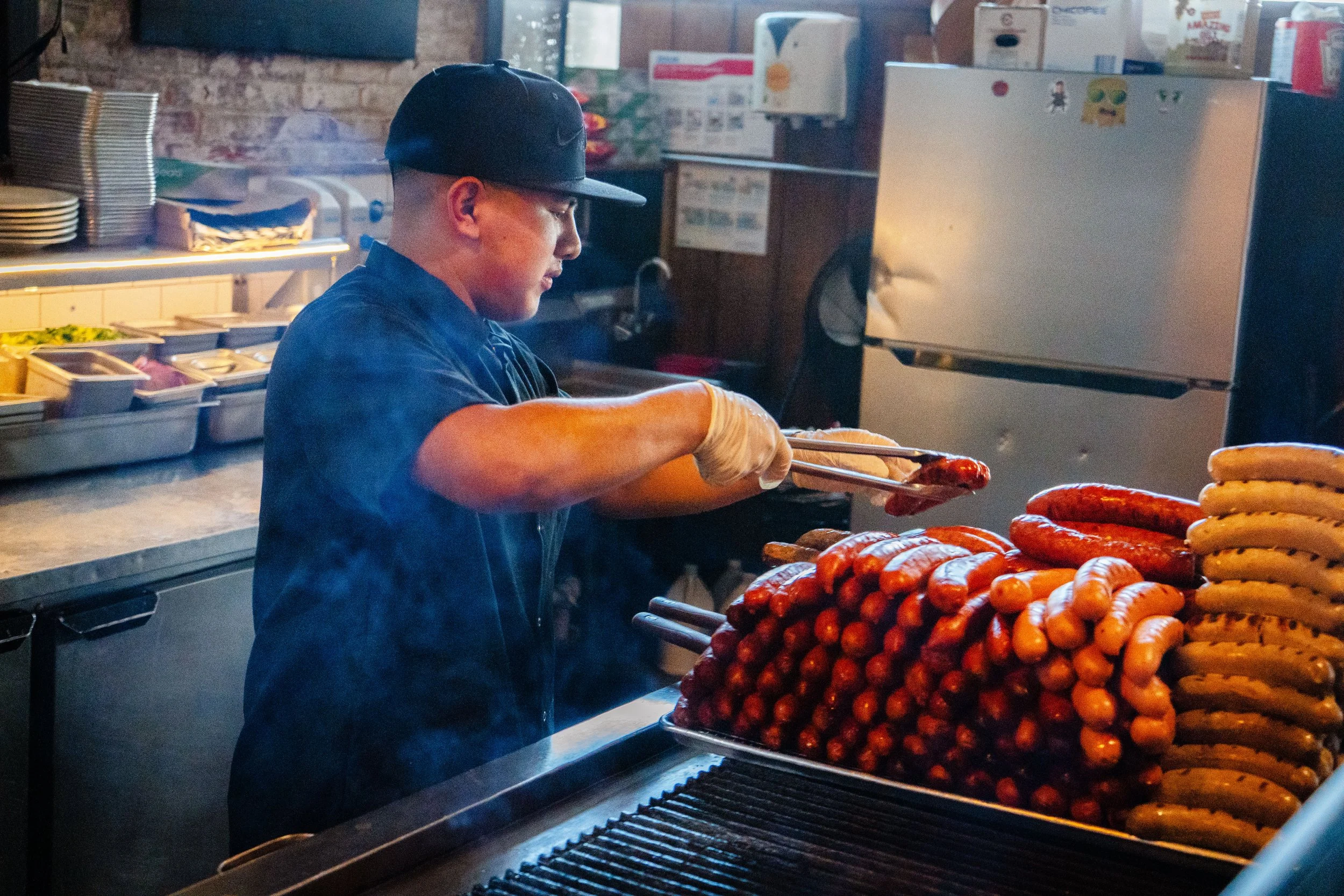 Person grilling sausages indoors, using tongs, stacks of sausages beside grill, wearing cap and gloves.