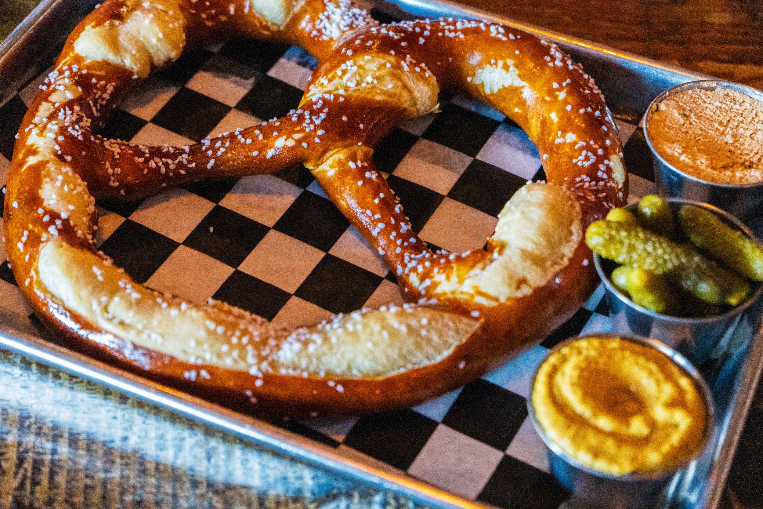 A large soft pretzel with coarse salt, served on a checkered paper-lined tray with mustard, pickles, and a cheese dip in small cups.