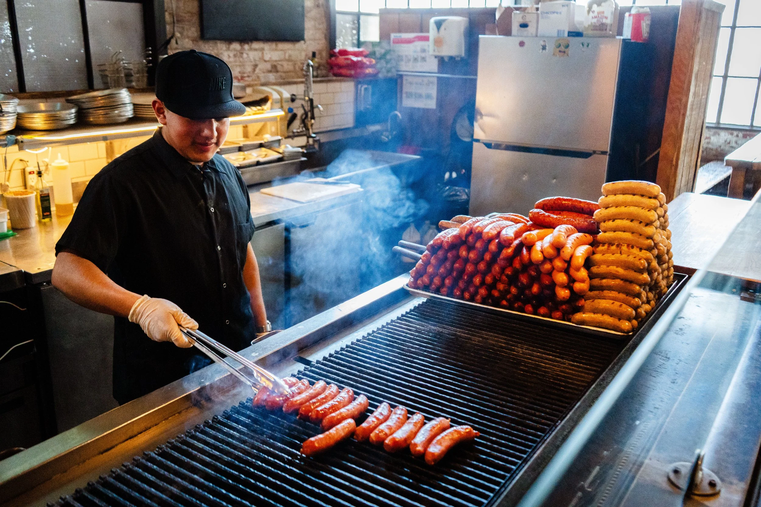 Chef grilling sausages in a restaurant kitchen with stacks of sausages on a tray nearby.