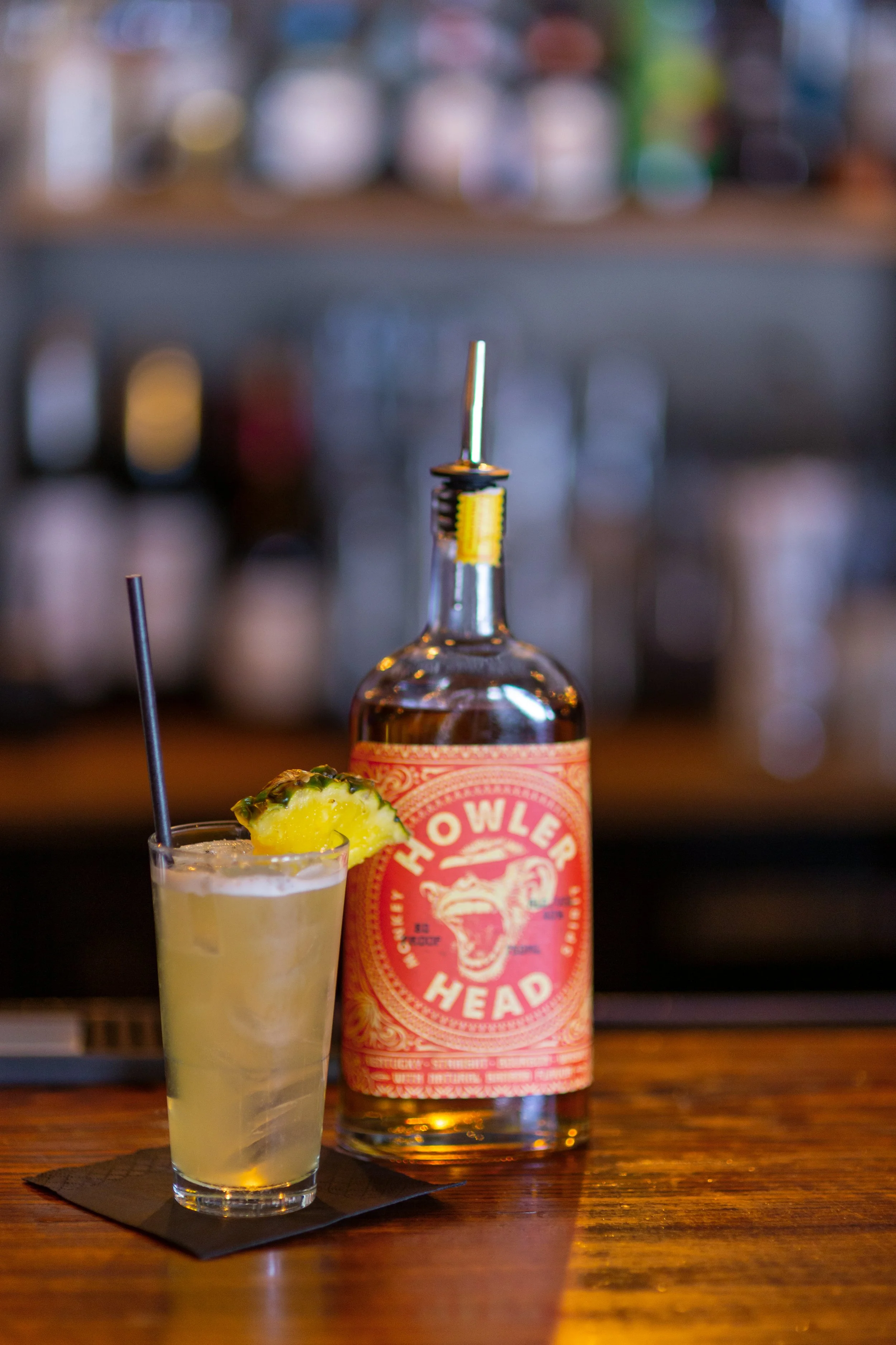 Cocktail in a glass with straw and pineapple garnish next to a Howler Head whiskey bottle on a bar counter.