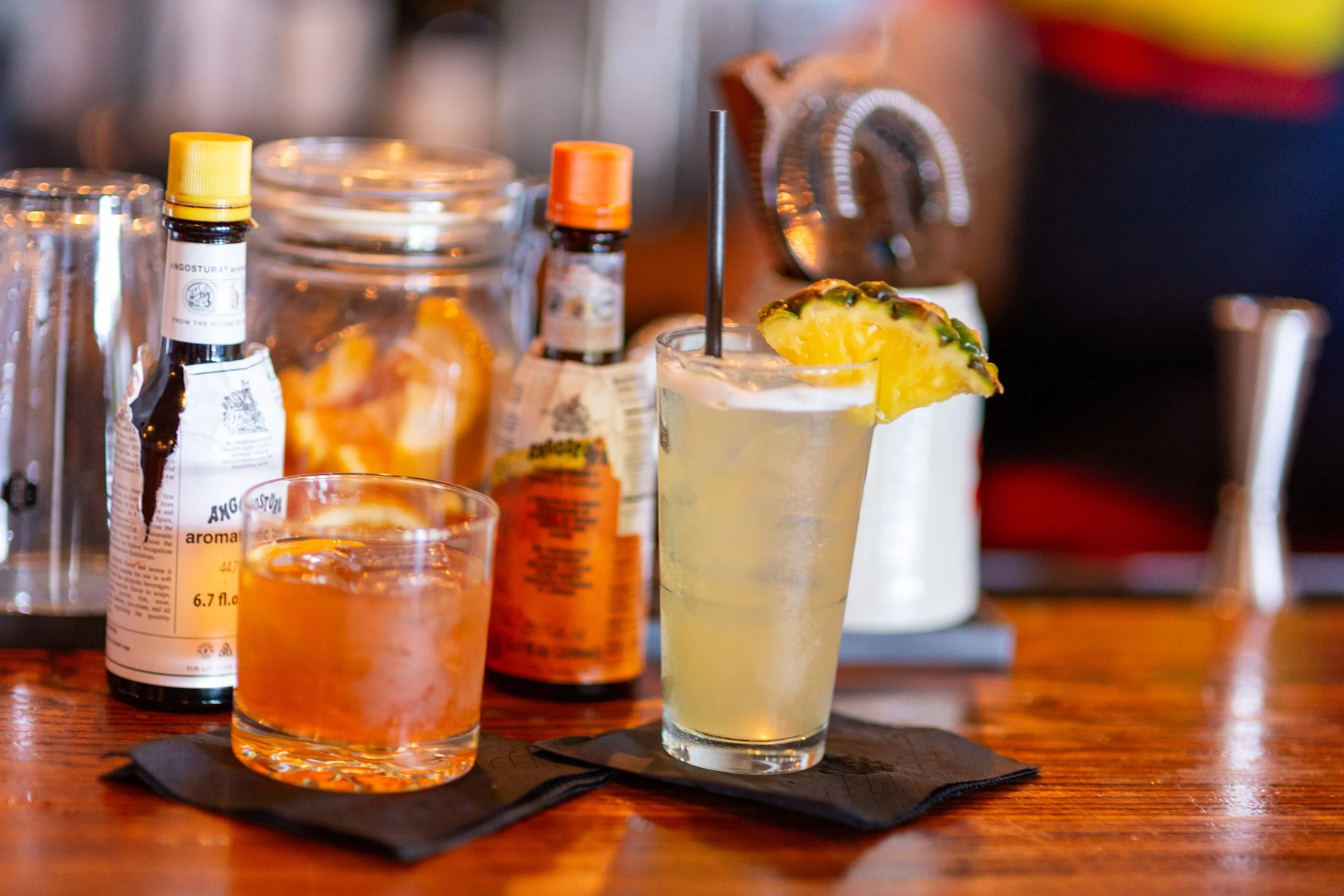 Two cocktails on a bar with bitters bottles and a jar of orange peels in the background.