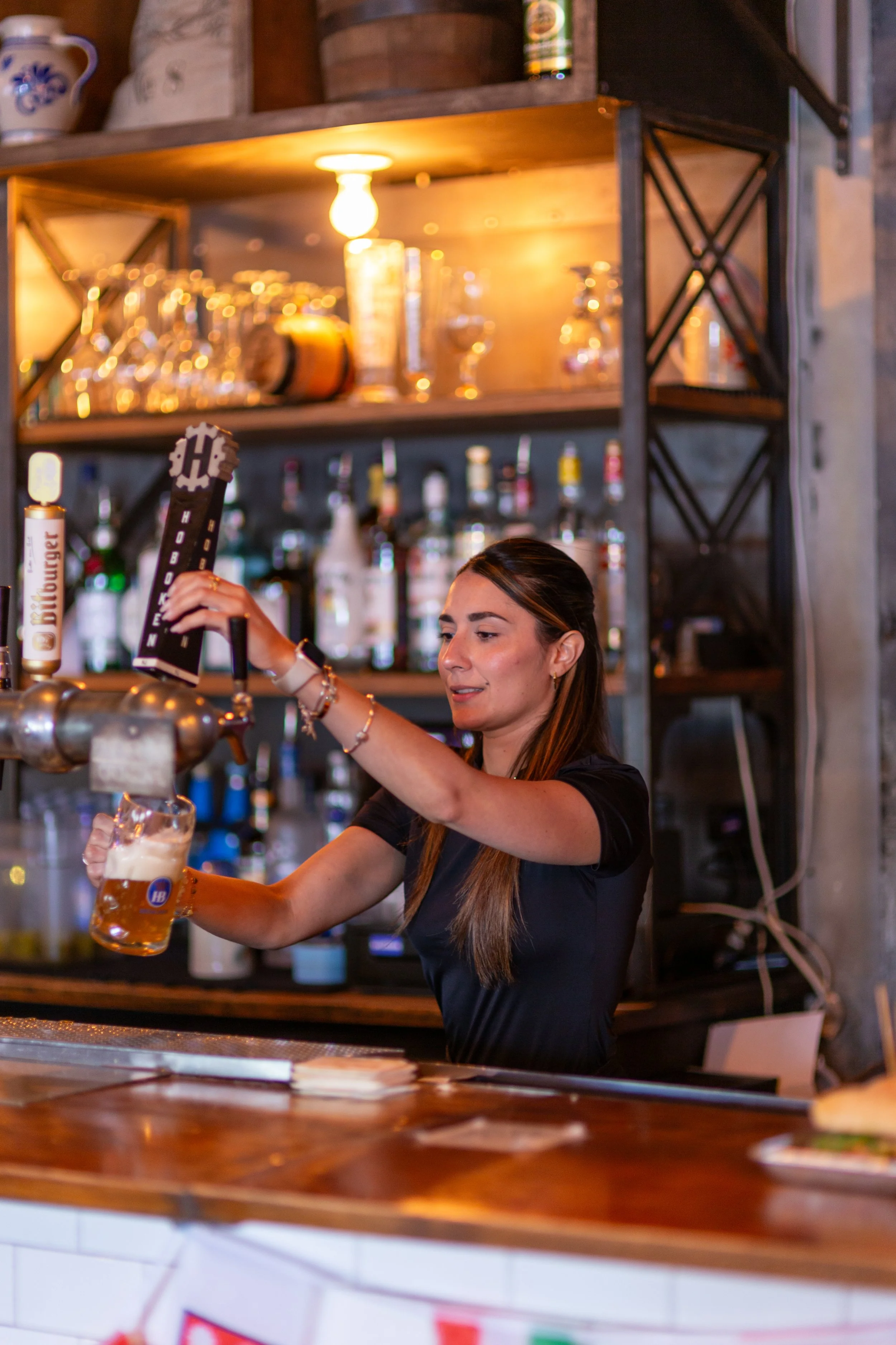 Bartender pouring beer from a tap into a mug at a bar, with shelves of glasses and bottles in the background.