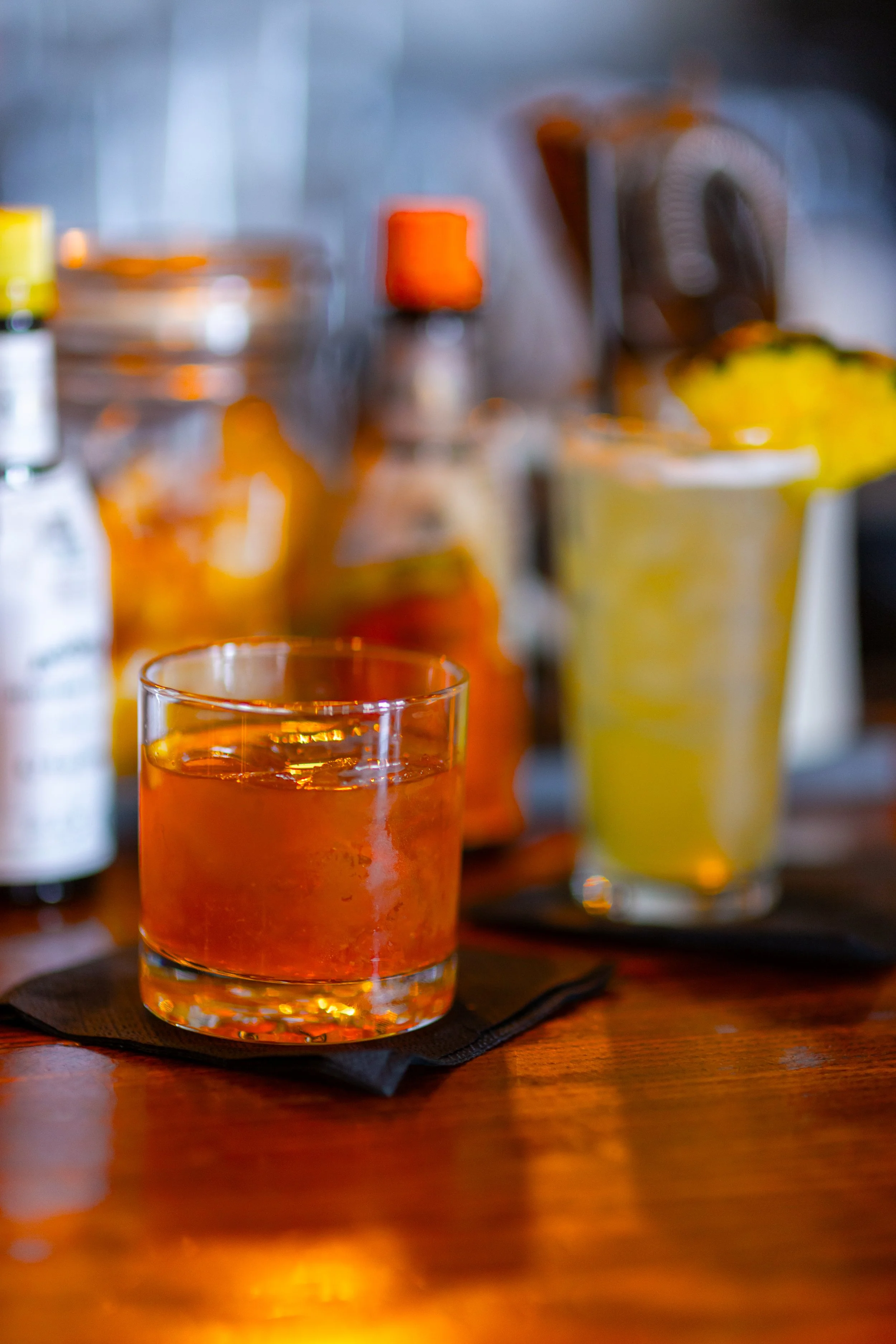 Close-up of a glass of whiskey on a wooden bar counter with a yellow cocktail in the background.