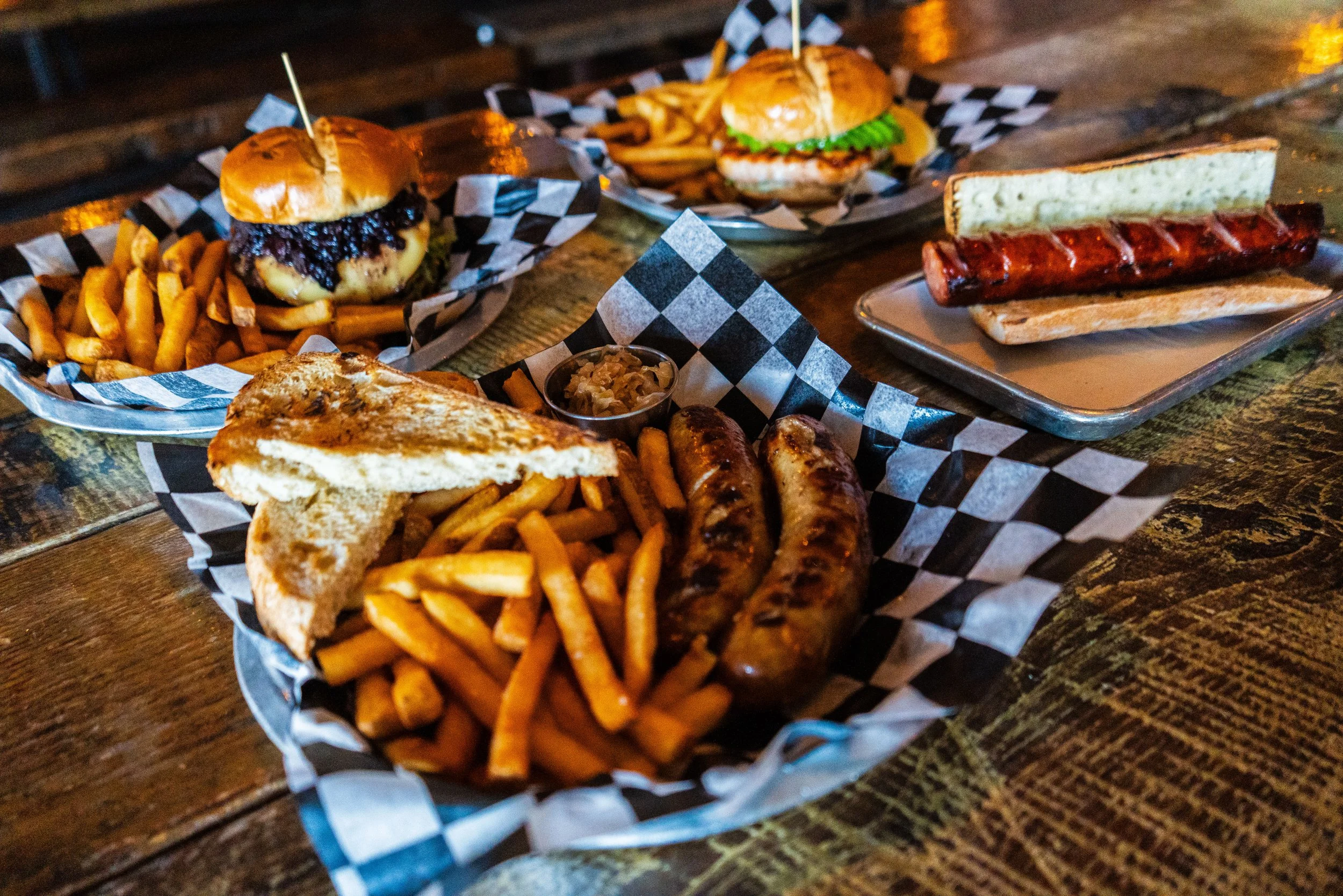 Assorted restaurant meals on a wooden table, including a burger with fries, another burger with lettuce, a hot dog on a bun, and a plate with sausages, toast, and fries.