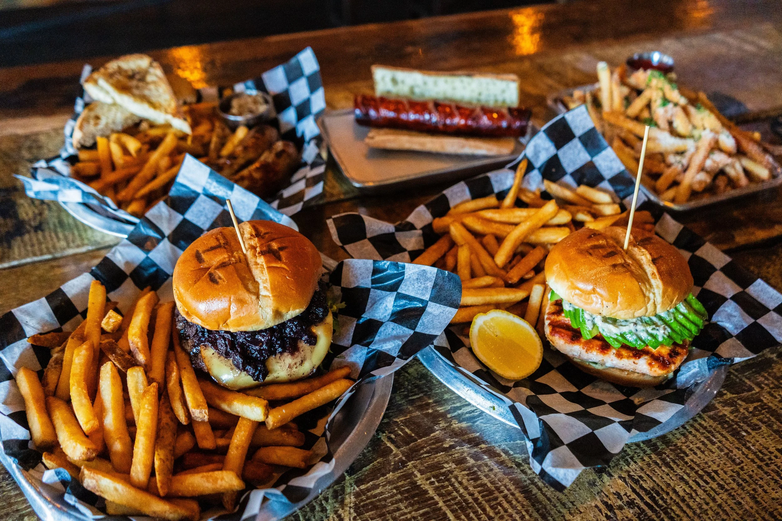 Plates of burgers with fries, a sausage on a bun, and a lemon slice on wooden table at a restaurant.