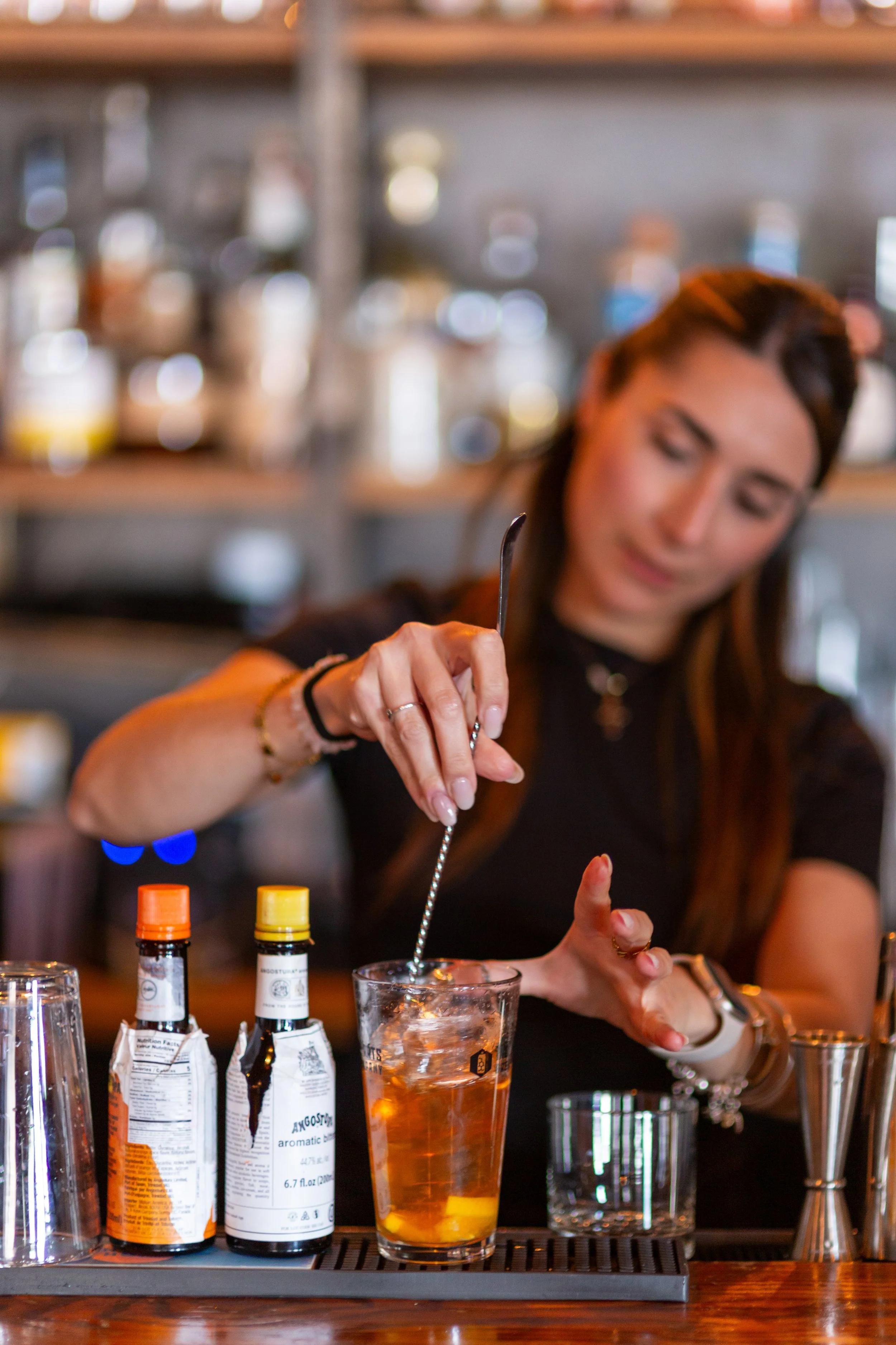 Bartender stirring a cocktail in a glass with ice, surrounded by bottles of Angostura bitters and a metal jigger on a bar counter.