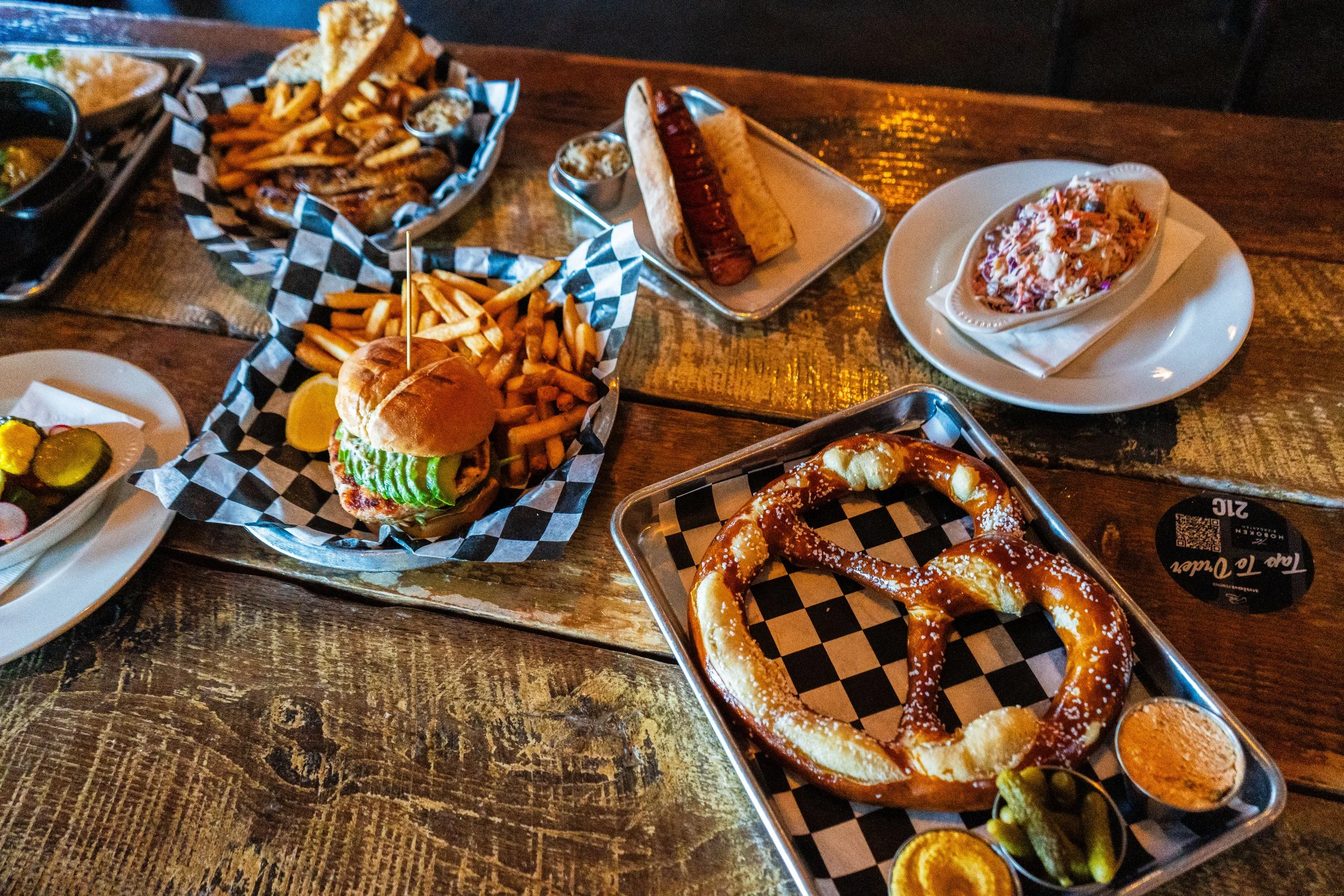 A variety of foods on a wooden table including a cheeseburger with fries, a pretzel with dipping sauces, a hot dog, a side of coleslaw, and a tray with sliced pickles and radishes.