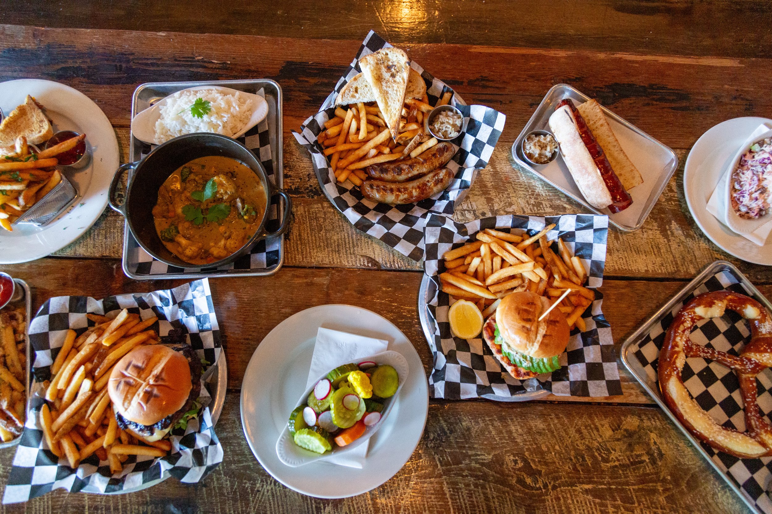 Assortment of dishes on a wooden table including burgers with fries, sausages, a hot dog, a curry with rice, pickled vegetables, and a pretzel.