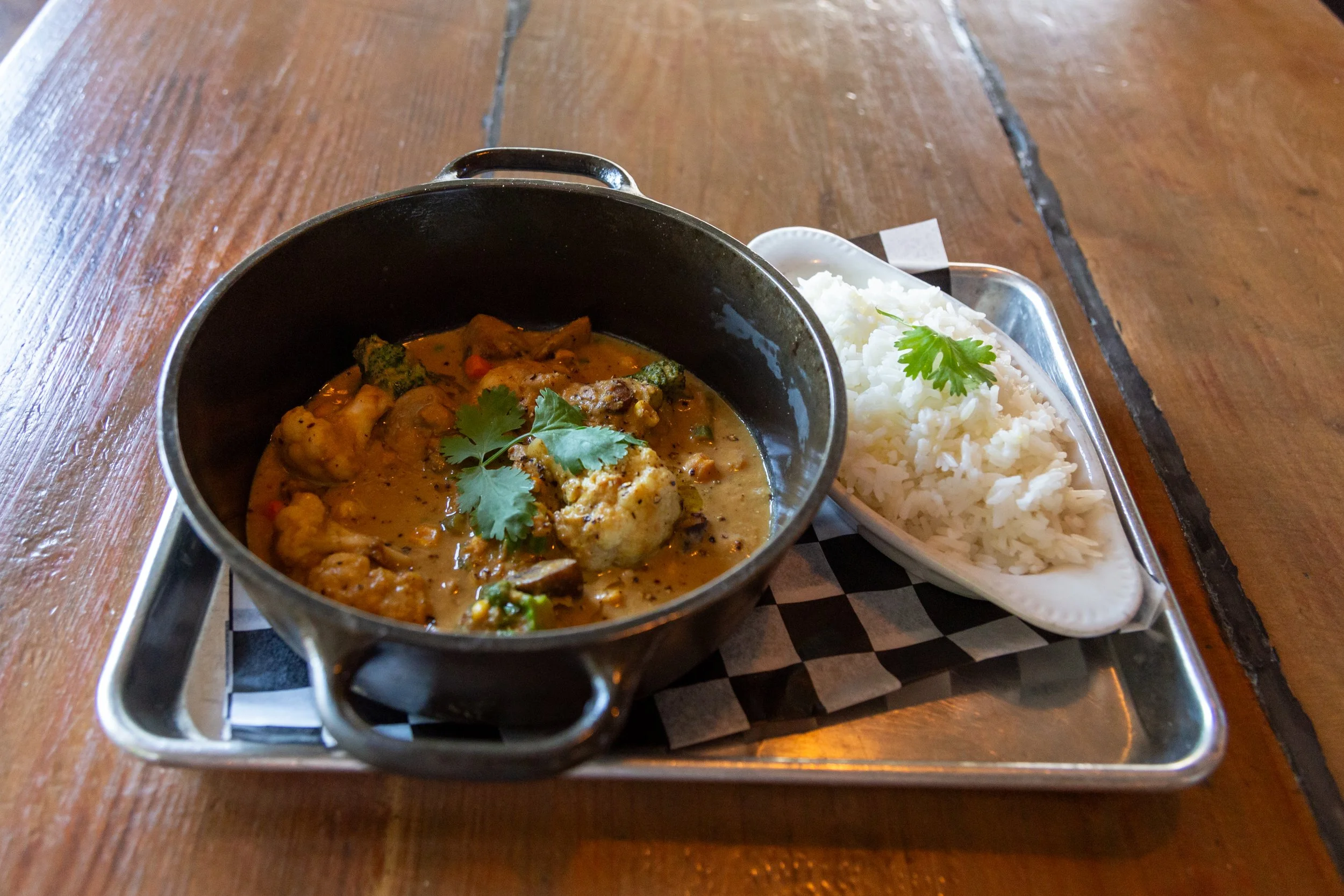 Plate with vegetable curry in a black bowl and a side of white rice garnished with cilantro, served on a wooden table.