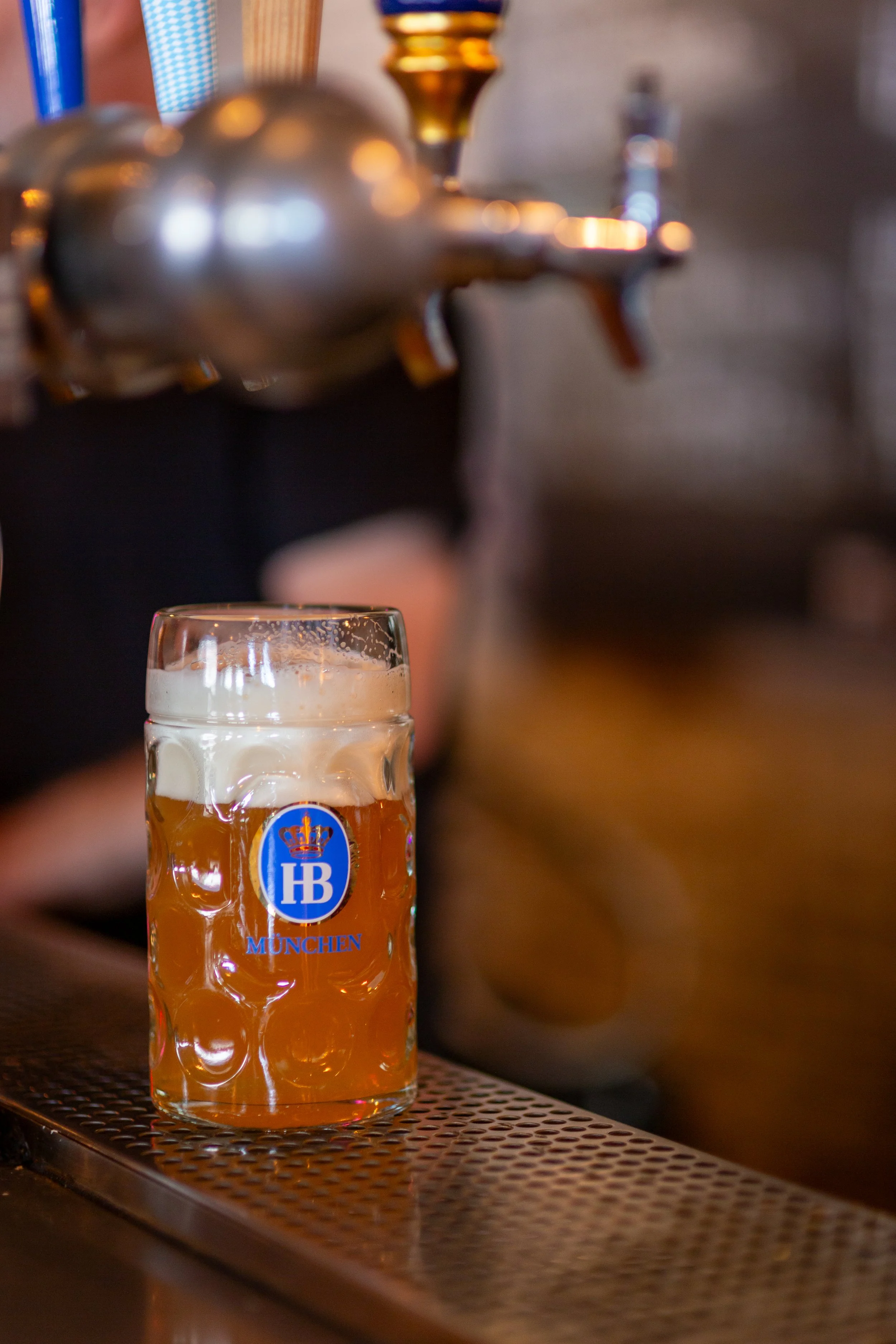 Glass of beer with foam under a tap, on a bar counter, labeled with "HB München."