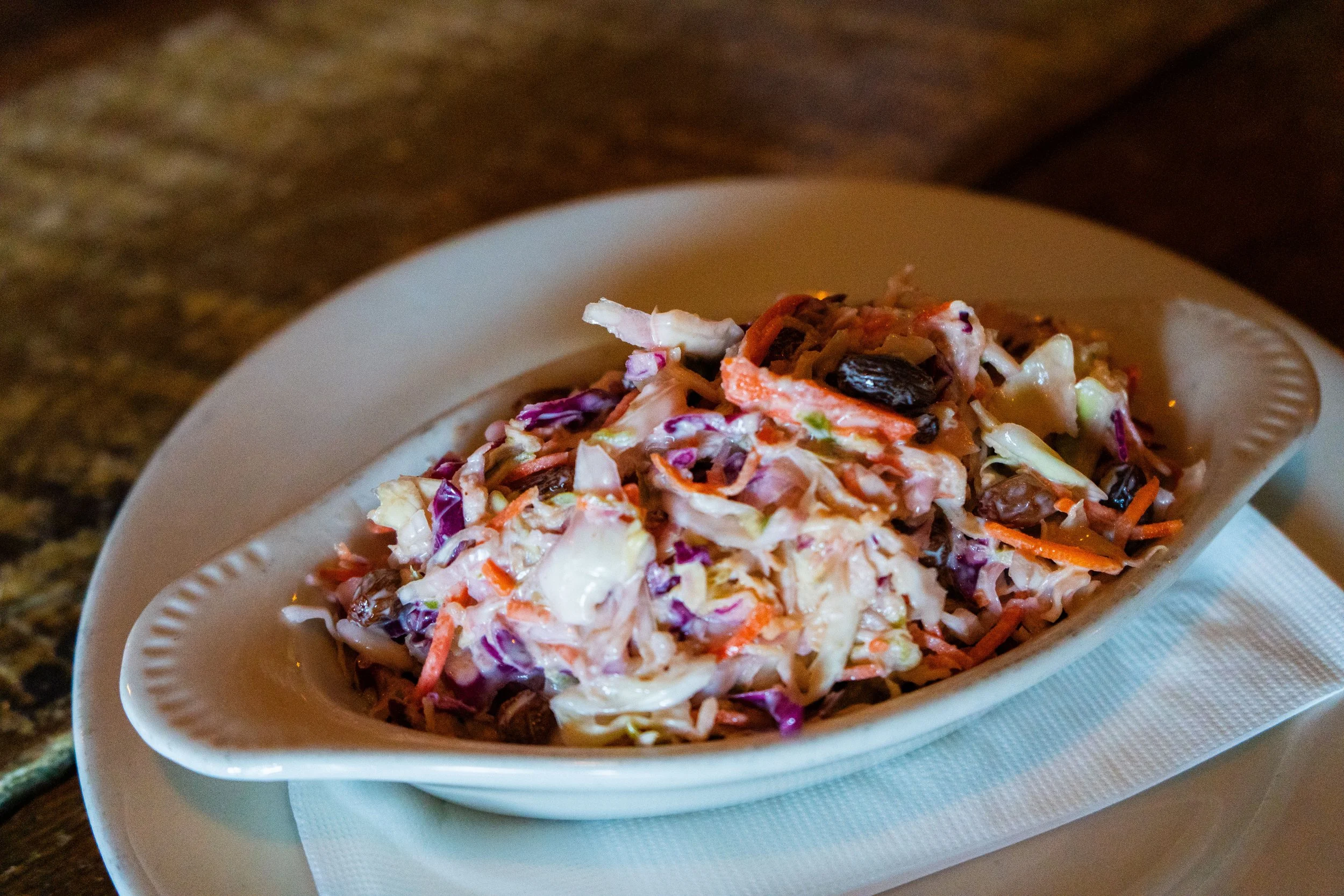 A dish of creamy coleslaw with cabbage, carrots, raisins, and a mayonnaise-based dressing on a white plate.