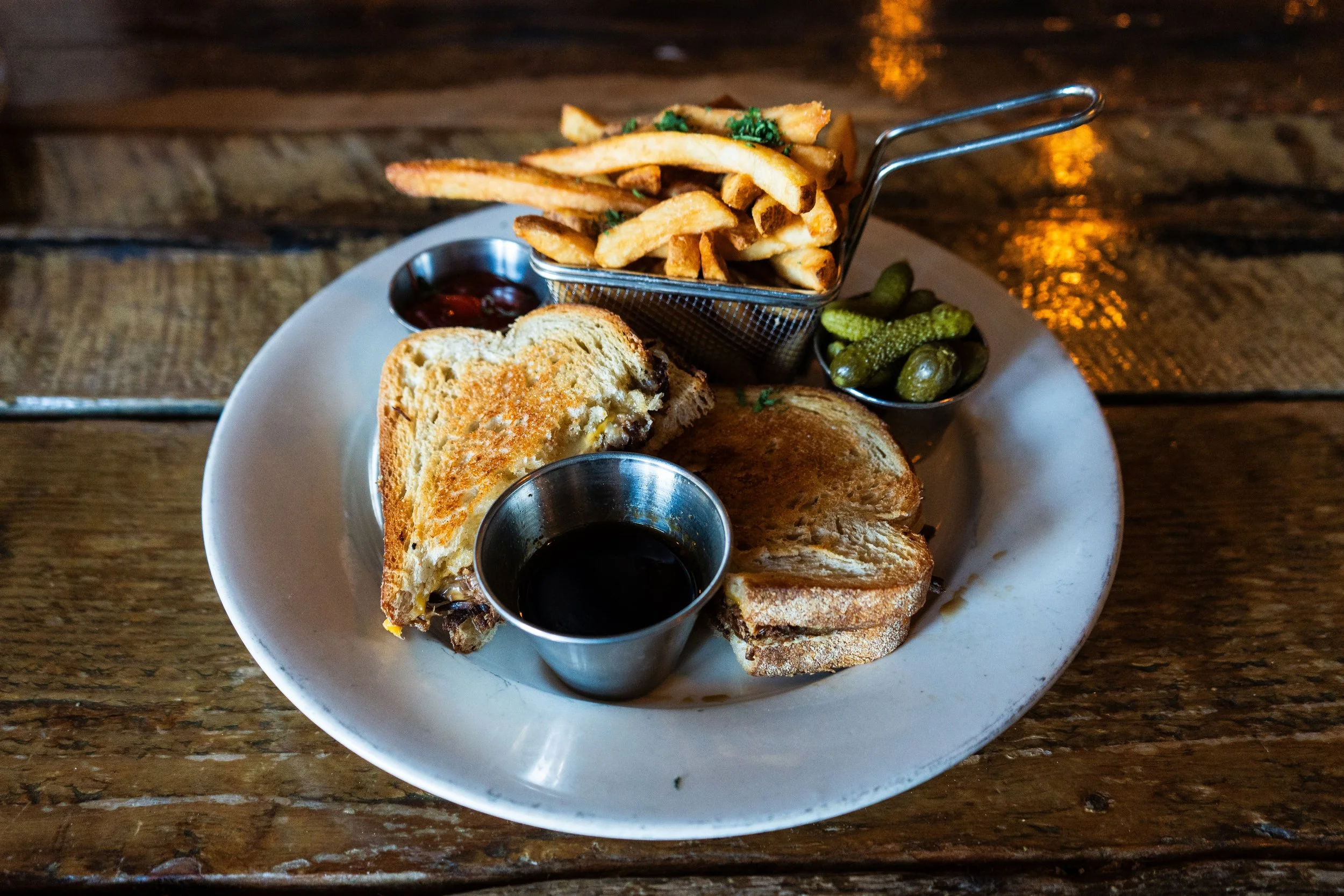 Plate with a grilled sandwich, fries in a metal basket, pickles, and dipping sauces on a wooden table.