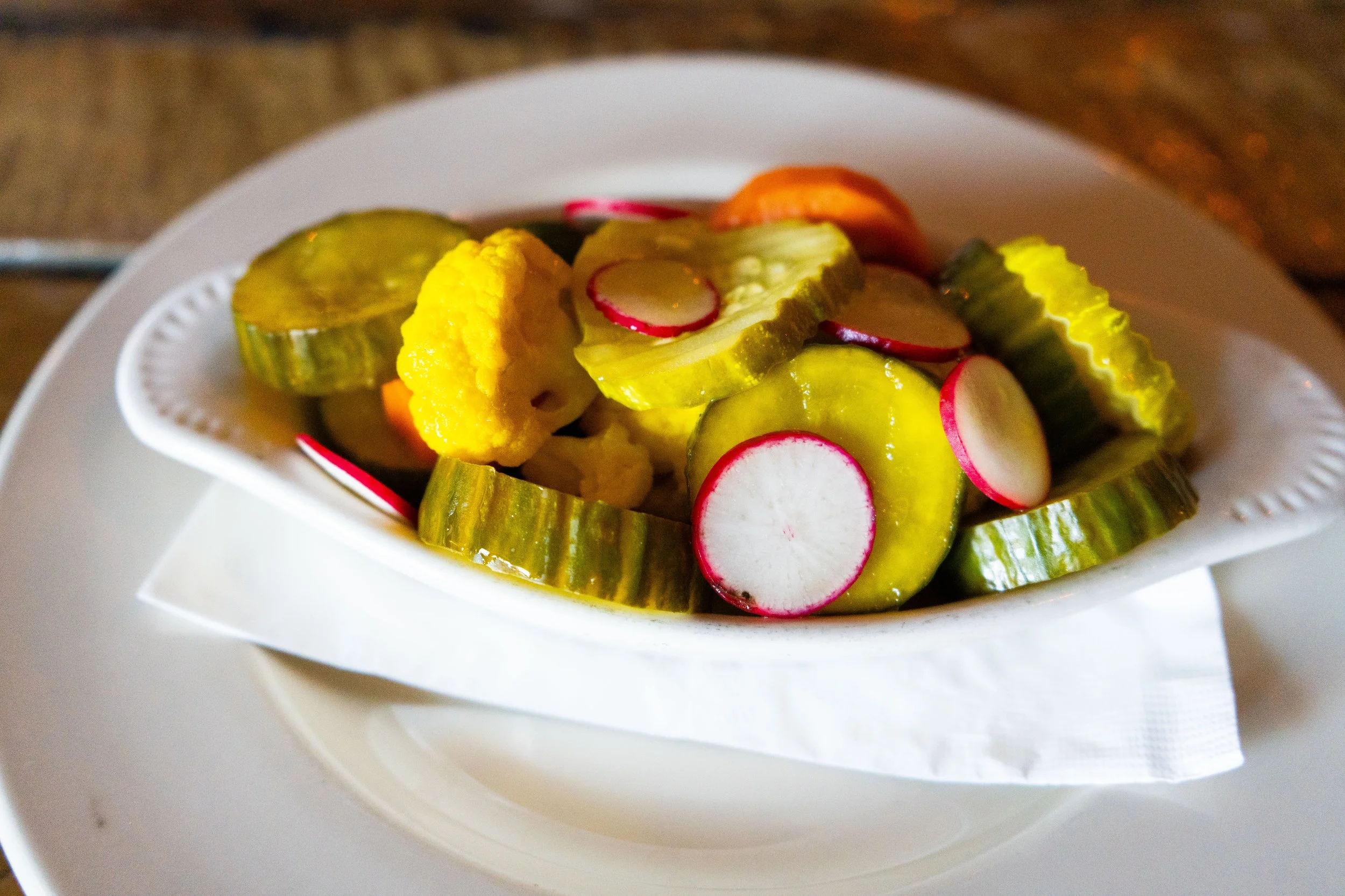 Assorted pickled vegetables in a white dish, including cucumber slices, radishes, cauliflower, and carrots, served on a white plate.
