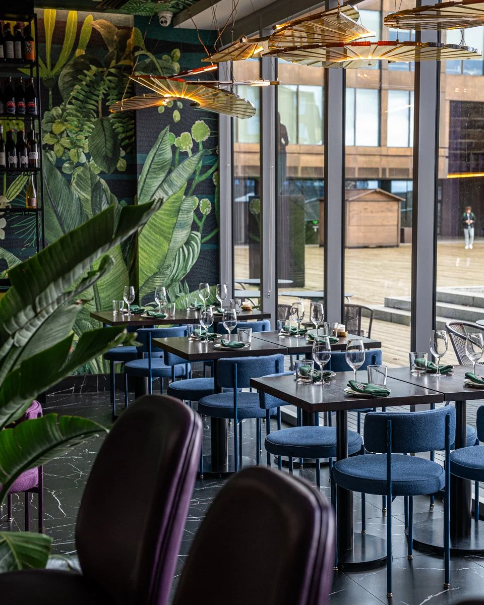 Restaurant interior with blue chairs at wood tables near large windows with plant mural on walls and overhead glowing chandeliers