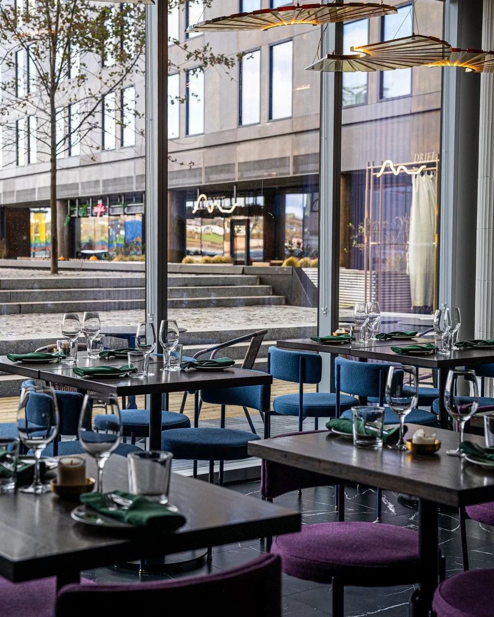 Restaurant interior with blue and purple chairs at wood tables near large windows with overhead glowing chandeliers