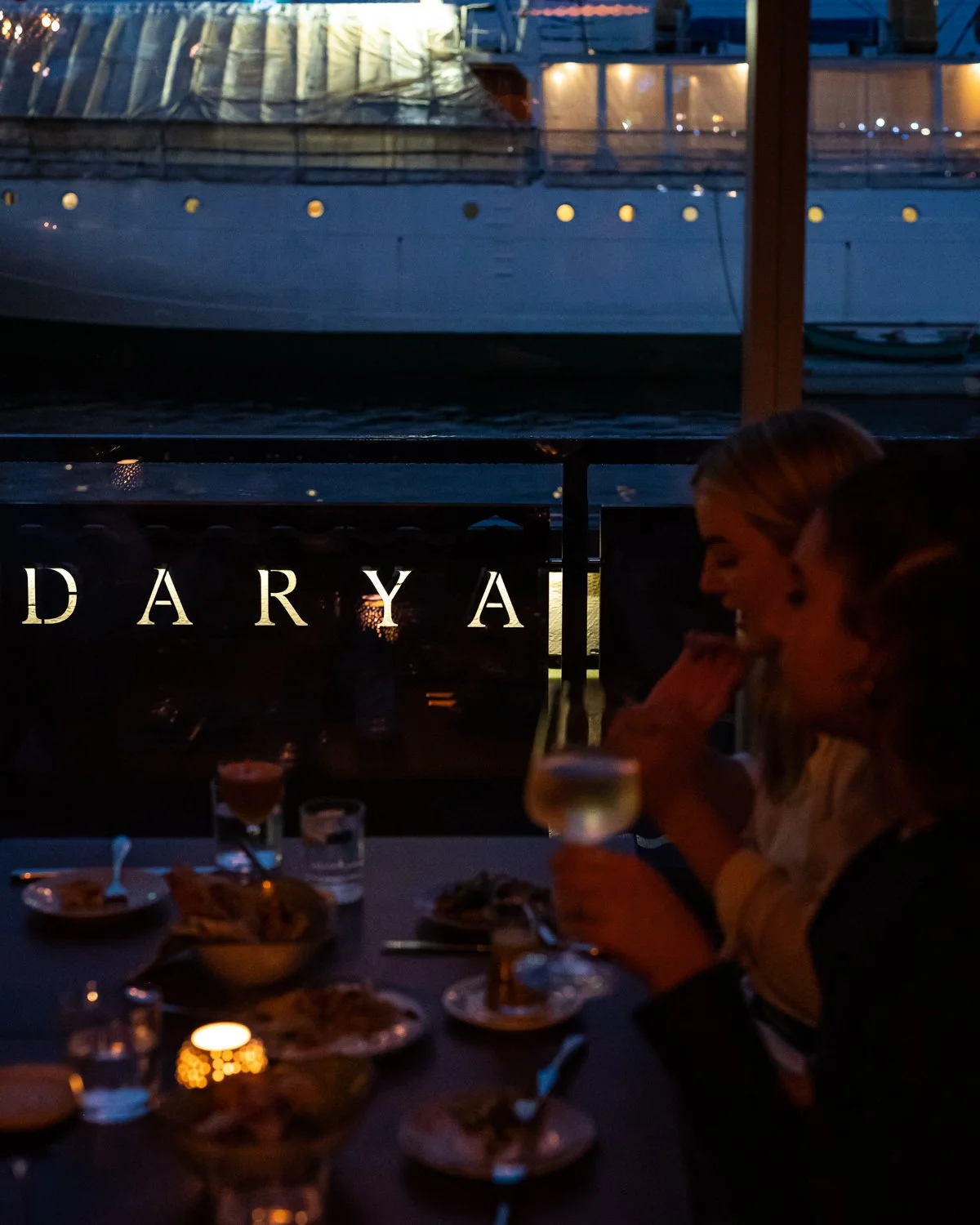 Restaurant guests dining on the patio at night with Darya signage on the railing and Halifax Harbour boats beyond