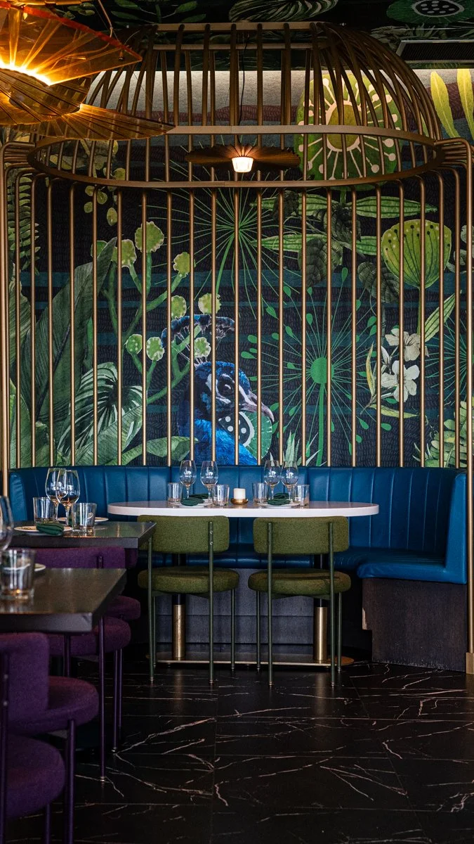 Restaurant interior with purple and green chairs, blue banquette, brass bird cage surrounding feature with peacock mural on wall